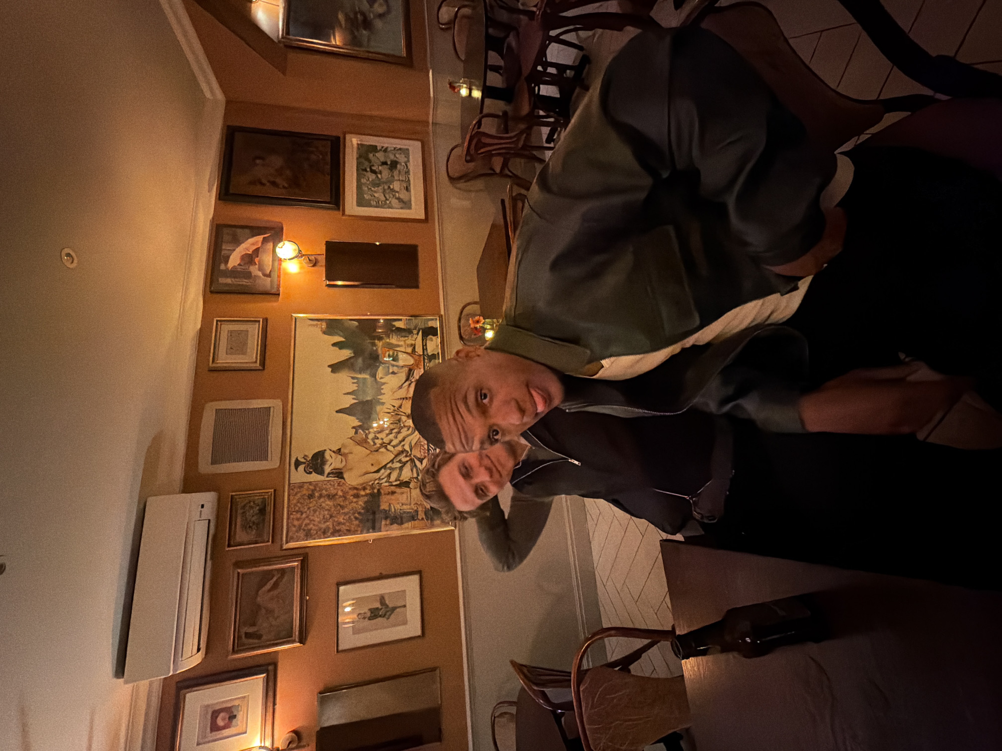 Two people are posing for a selfie in a room with multiple framed pictures on the wall. The person closest to the camera is sticking out their tongue. There is a table in the foreground with a wine bottle on it. The location appears to be a restaurant or pub. It is not possible to confirm the city with only this picture.