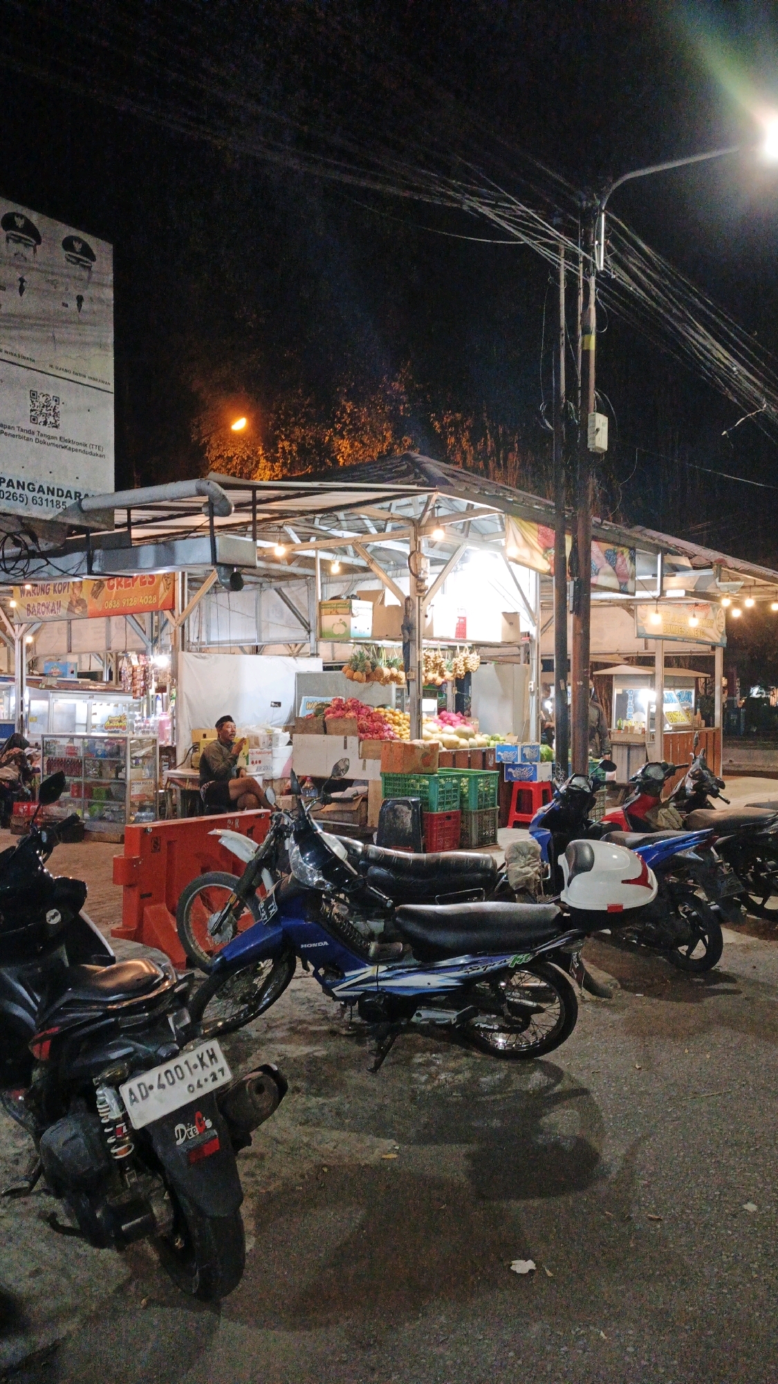 At night in Desa Karangbenda, Indonesia, a street-side market stall is illuminated by overhead lights. The stall displays a variety of fruits, including pineapples, what appear to be watermelons, and other produce. Crates of fruits are stacked, some colored green and red. Behind the produce, a person is seated at a table, eating. Several motorcycles are parked in front of the stall on an asphalt surface. One motorcycle in the foreground has a license plate reading "AD 4001 KH". The background shows more market stalls and a large sign with text and images, suggesting a public notice or advertisement. Electrical wires and a utility pole are visible overhead, contributing to the urban street environment.