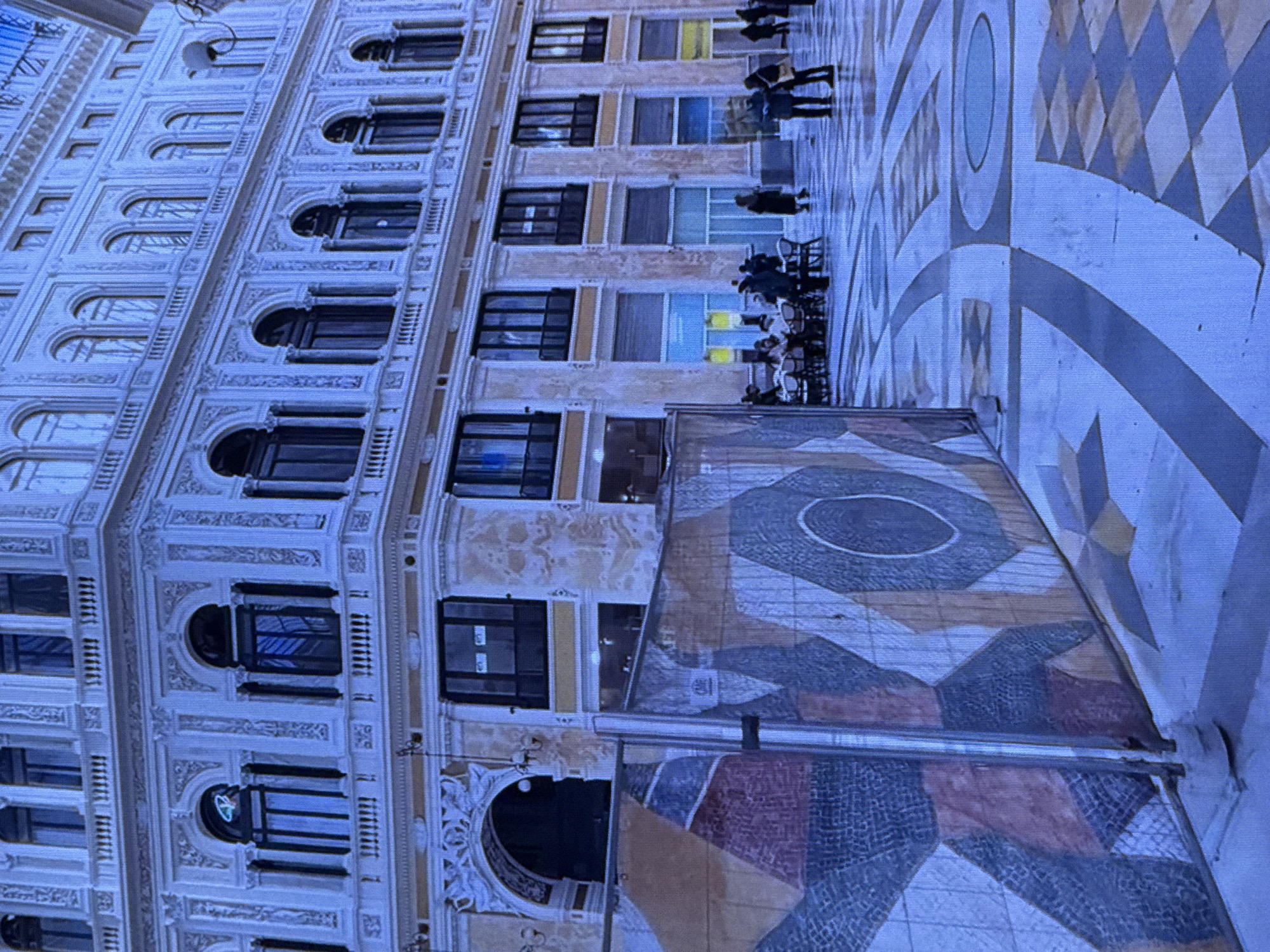 The image depicts an outdoor plaza in front of an ornate, multi-story building with classical architectural elements. The building is light-colored with numerous arched windows, balconies, and decorative moldings. In the foreground, a construction barrier with a colorful mural is visible, partially obscuring the view of the plaza. The plaza floor has a geometric pattern of inlaid stones. Several people are present, some seated at tables outside what appears to be a cafe, while others are standing and walking. The lighting suggests it is daytime, possibly overcast. This scene is located in the Piazza del Plebiscito in Naples, Italy.