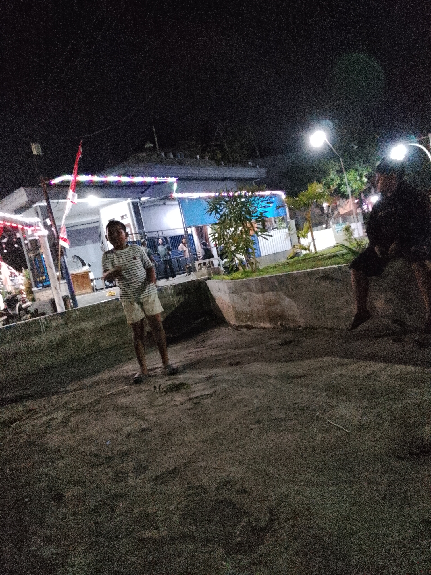 The image depicts an outdoor scene at night, likely in a public area within the city of Pasuruan, Indonesia. In the foreground, a young boy stands facing the camera, wearing a striped t-shirt and shorts. His hands are partially obscured, suggesting he might be in motion or holding something. Behind him, to the left, is a building illuminated by string lights and possibly a red and white Indonesian flag. Further in the background, other individuals can be seen, including one person seated on a low concrete wall on the right side of the image. The environment appears to be a paved area with some vegetation, suggesting it could be a park, a street, or an area adjacent to a commercial establishment. The lighting indicates it is nighttime, with streetlights providing illumination. The overall scene is somewhat grainy, possibly due to low light conditions.