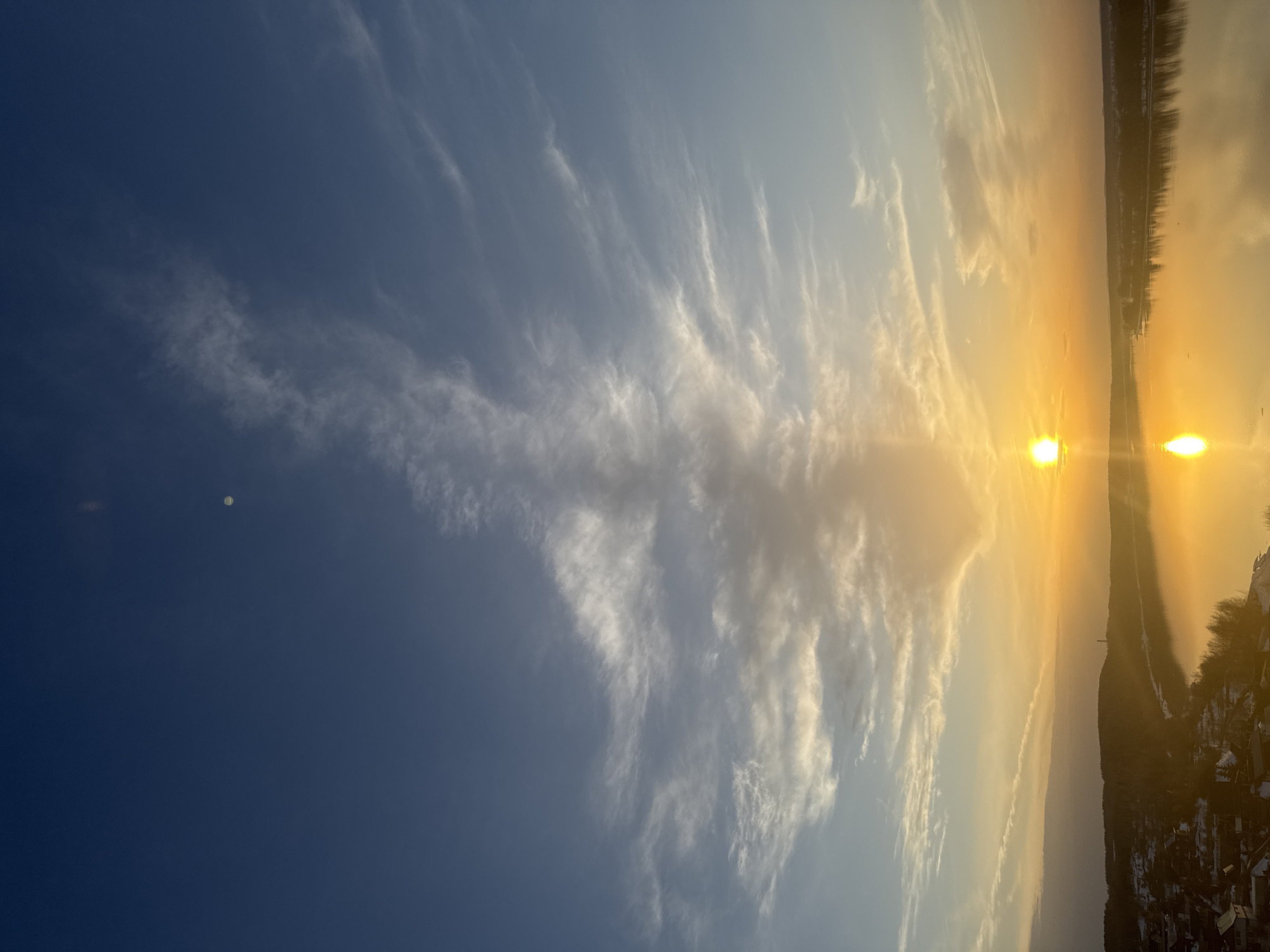 The image shows a sunset over a body of water, likely a river or lake, in Ольховка, Russia. The sun is positioned low on the horizon, creating a bright, golden reflection on the water's surface. The sky transitions from a deep blue on the left to a lighter, orange-tinged hue near the horizon where the sun is setting. Wispy, cirrus-like clouds are scattered across the sky, with a more prominent cluster of clouds closer to the setting sun.
The horizon line shows the silhouettes of trees and buildings, possibly indicating a shoreline or a developed area. Snow can be seen on some of the roofs of the houses along the horizon. The scene is still.