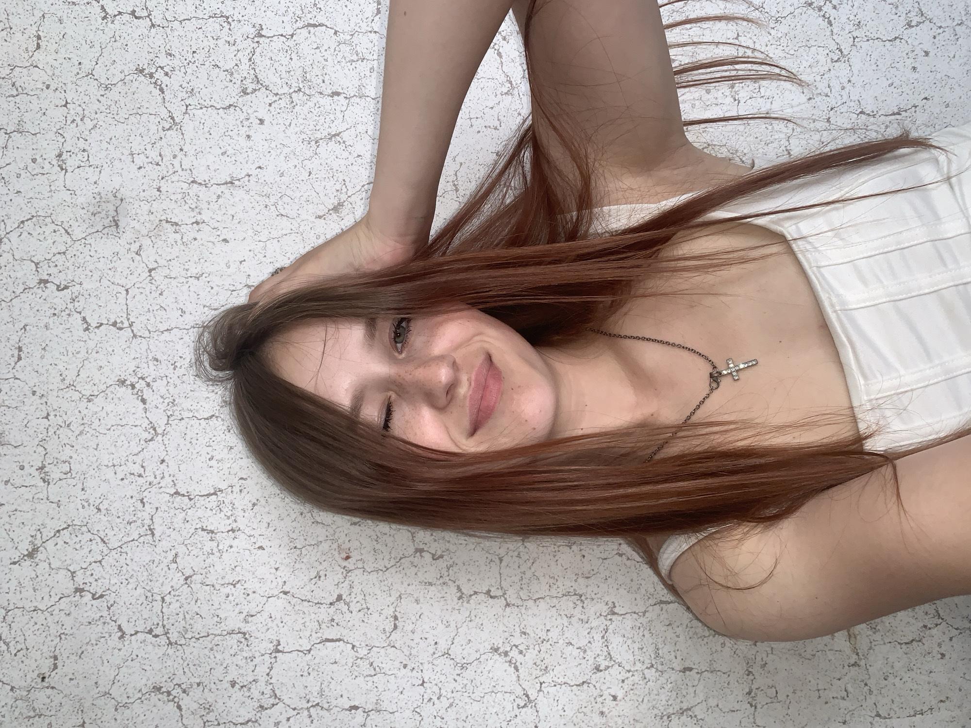 A young woman with long, reddish-brown hair is lying on a light-colored, cracked surface. She is wearing a white top and a necklace with a cross pendant. Her left hand is resting on her head. She is smiling and looking directly at the camera. The location of the image is unknown, there is no evidence that the image was taken in Фоки, Russia.