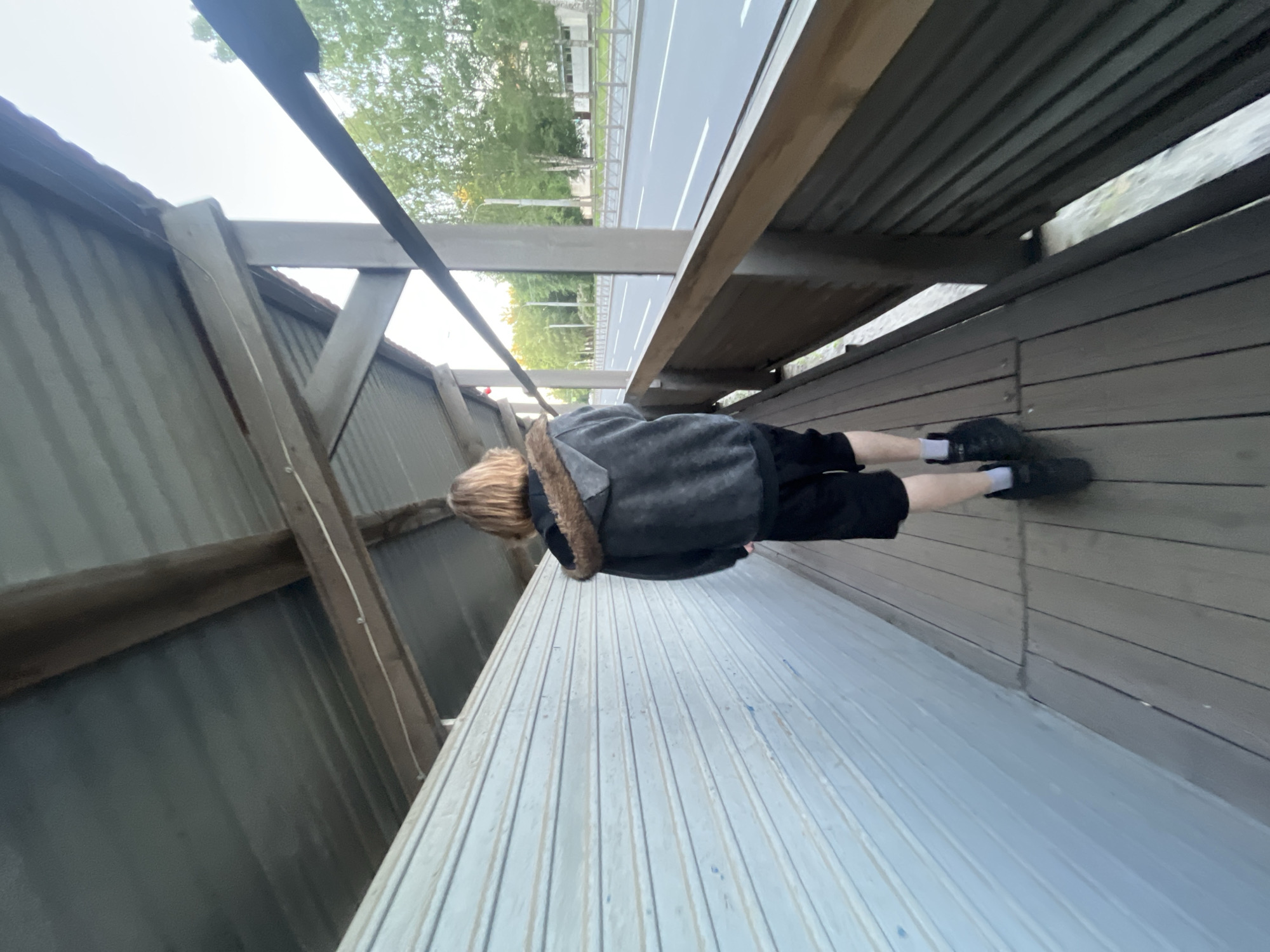 A person, seen from behind, walks along a covered pedestrian walkway in Saint Petersburg, Russia. The individual has shoulder-length light brown hair and wears a dark grey hooded sweatshirt with a brown faux-fur hood lining, black shorts, and dark sneakers with white ankle socks. The walkway floor consists of dark, horizontally laid wooden planks. To the left, a light blue corrugated metal wall with horizontal ribbing parallels the path. Overhead, a dark grey corrugated metal roof is supported by an exposed timber beam structure, with a thin white cable running along one beam. To the right, a wooden railing with horizontal slats separates the path from a multi-lane paved road. White lane markings are visible on the asphalt surface. Beyond the road, a median strip features mature deciduous trees, some with light-colored bark, and a low, light-colored fence parallels the distant roadside. The sky is bright and overcast.