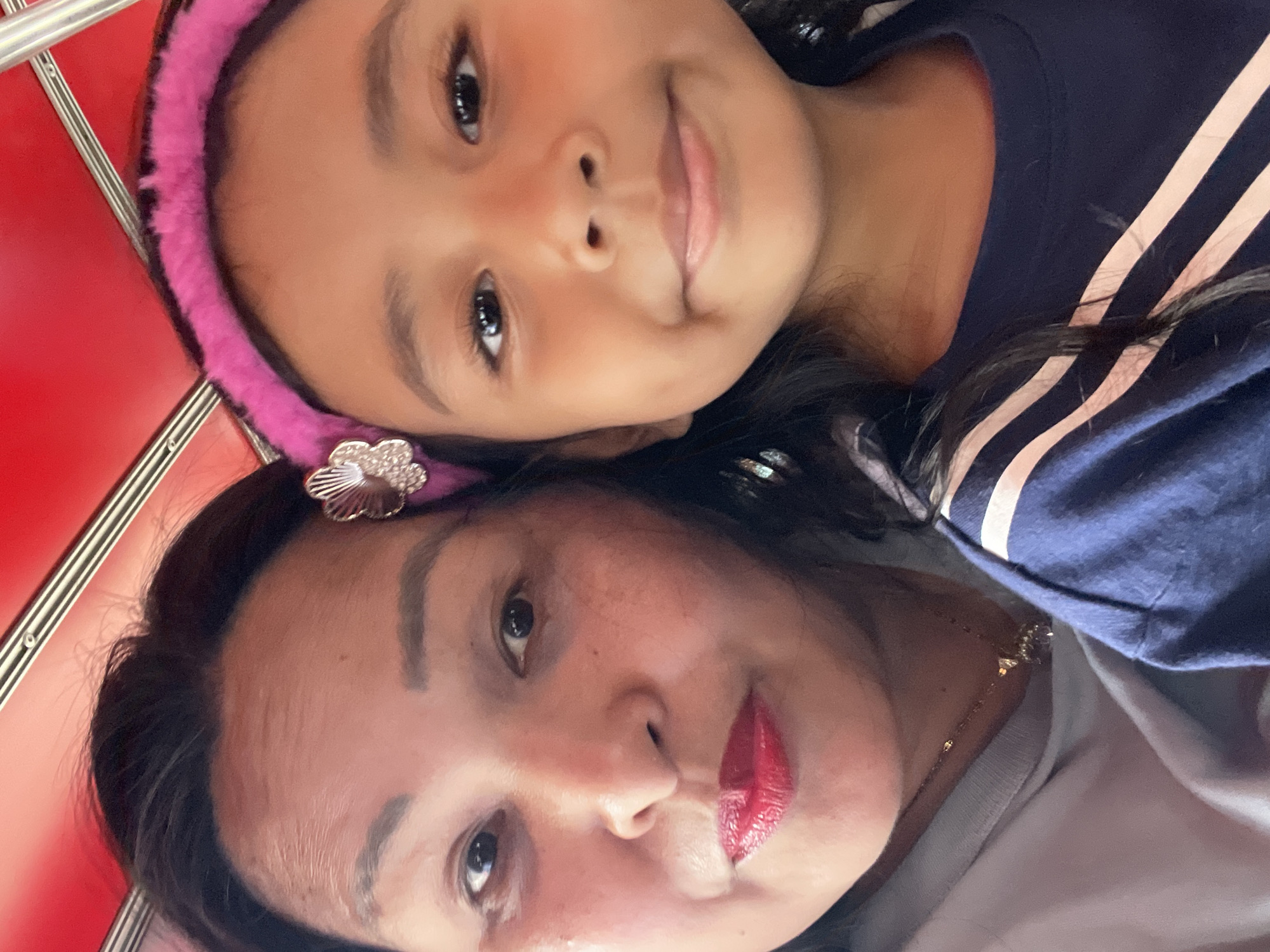 An adult woman and a young girl are depicted in a close-up selfie within the interior of a vehicle, characterized by a vibrant red background with horizontal metallic railings. This background is consistent with the passenger cabin of a tricycle, a common mode of transport in Davao City, Philippines.
The woman, on the left, is presumed to be middle-aged. She has dark hair, styled back, and wears a light-colored, floral hair clip with clear stones above her right ear. Her lips are coated with red lipstick, and she wears a fine gold-colored chain necklace with a dark pendant over a light grey top. Her gaze is directed at the camera with a slight smirk.
The girl, on the right, appears to be approximately 5-8 years old. She has dark hair and wears a fluffy pink headband. Her expression is a gentle smile, and she is wearing a navy blue t-shirt adorned with three light pink stripes on the shoulder. Her gaze is also directed at the camera. Both individuals are positioned closely together, indicating a shared photographic moment.