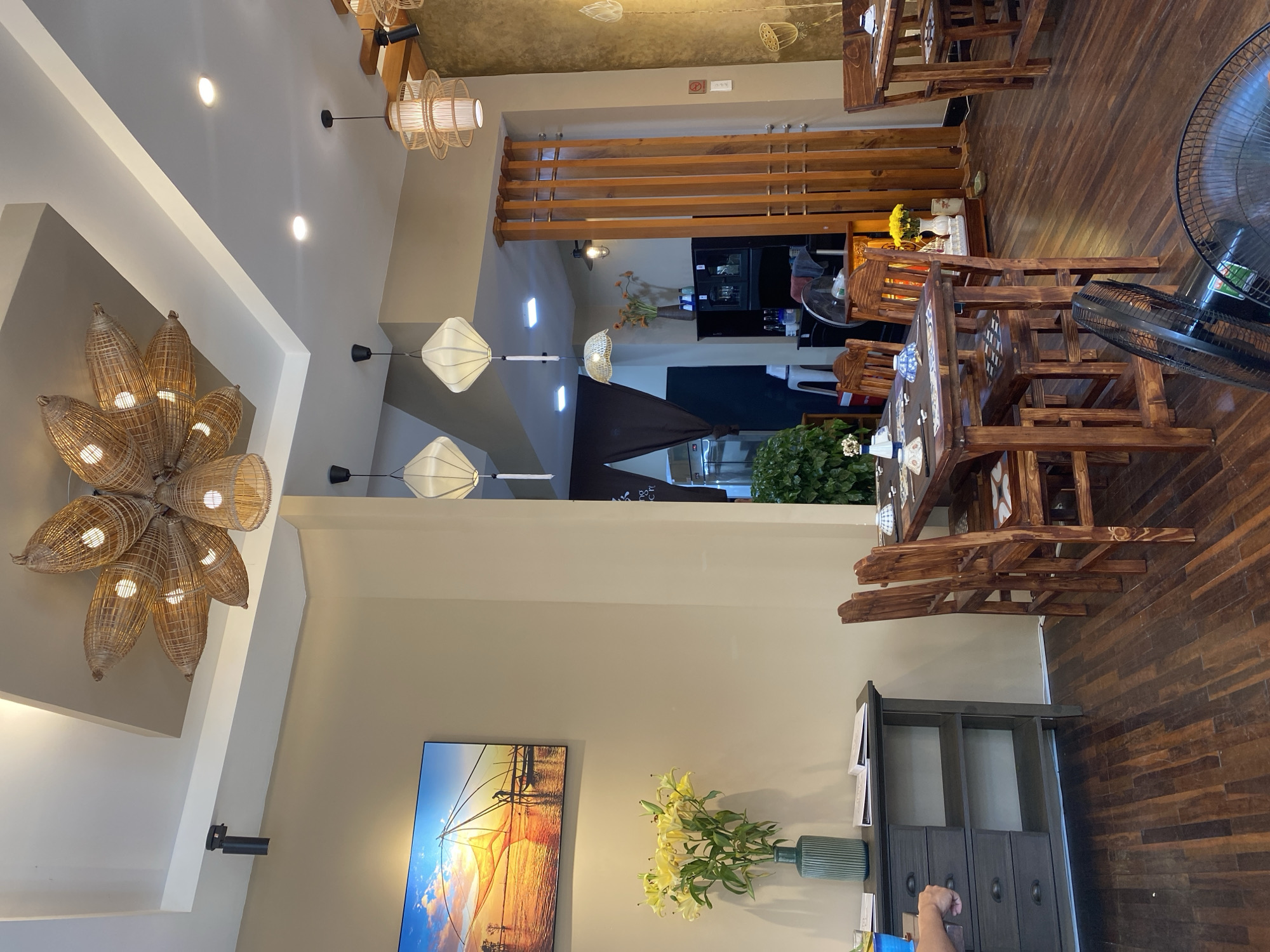 This image depicts the interior of a restaurant in Da Nang, Vietnam, with a focus on its decor and ambiance. The restaurant features a warm and inviting atmosphere with wooden furniture and natural materials.
In the foreground, a dark wood table is set with plates and bowls, suggesting a dining setting. Several wooden chairs surround the table. A dark grey cabinet with drawers and shelves is positioned to the left of the table. A tall green vase holding a bouquet of yellow lilies sits on top of the cabinet. To the left of the cabinet, a hand is visible reaching towards a drawer.
The ceiling is a prominent feature, with a unique woven rattan light fixture shaped like a flower. Other pendant lights with woven shades and square lantern-style lights hang from the ceiling, adding to the restaurant's rustic charm.
The walls are painted in a neutral beige color. A large framed picture of a sunset scene over water with fishing nets hangs on the left wall.
In the background, more dining tables and chairs are visible. A wooden divider with vertical slats separates different areas of the restaurant. A potted plant with lush green leaves adds a touch of nature to the space. The flooring is made of dark wooden planks.
The overall impression is of a cozy and aesthetically pleasing restaurant, likely serving Vietnamese cuisine, with a strong emphasis on natural textures and warm lighting.