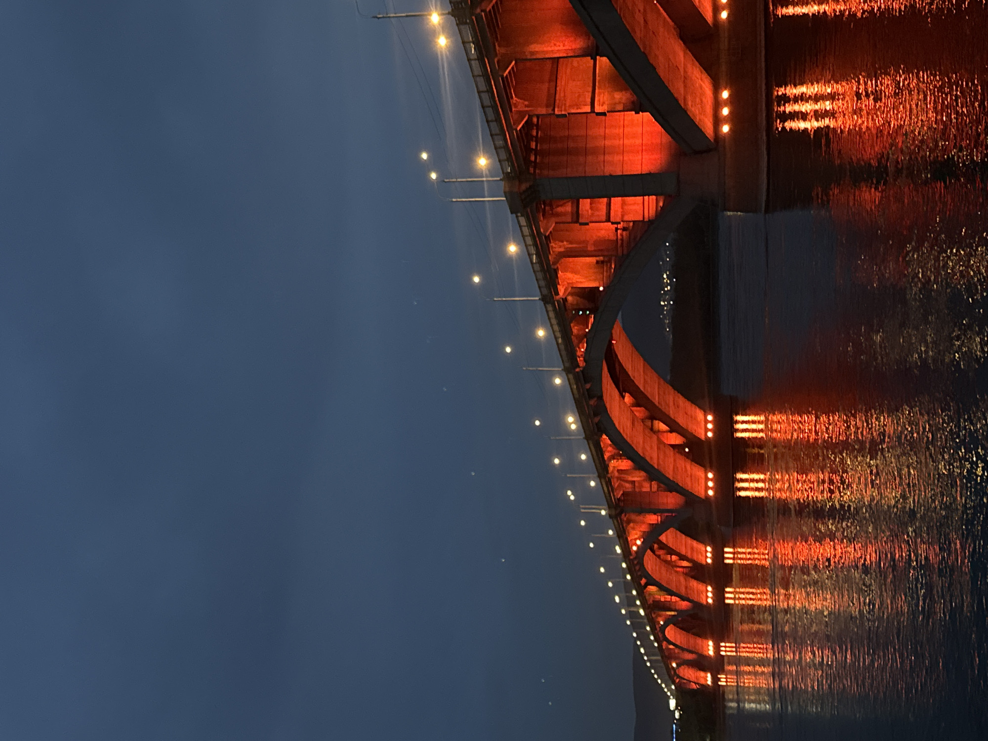 The image shows a large bridge illuminated with red lights, spanning a dark body of water. The bridge has multiple arched sections and is lined with streetlights. The water reflects the red illumination from the bridge, creating a vibrant glow. The sky above is a deep blue, suggesting it is either dusk or night. In the distance, faint lights can be seen along the shoreline, indicating a city. This is the Communal Bridge in Krasnoyarsk, Russia, a prominent landmark that crosses the Yenisei River.