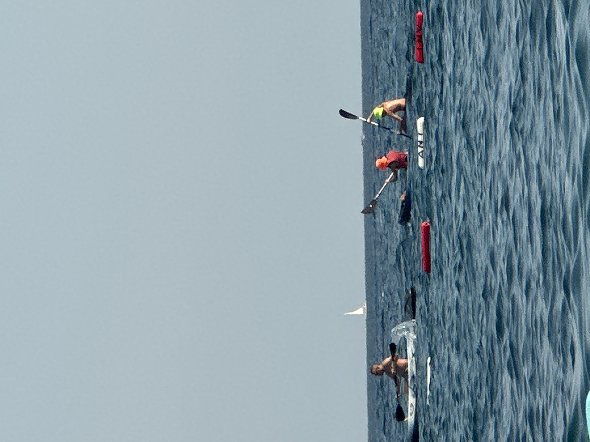 In the waters off the coast of Boreti, Montenegro, three individuals are engaged in kayaking. The scene captures a clear, bright day with a calm, blue sea under a pale blue sky. In the foreground, the choppy waves suggest gentle movement of the water.
To the left, a person in a kayak is paddling, with their torso visible above the waterline. Their kayak is white. Further to the right, two individuals are also in kayaks. One person, wearing a red life vest and an orange hat, is actively paddling. Beside them, another person, wearing a yellow hat, is also paddling. Both of these kayaks appear to be dark in color.
Several buoys are visible in the water, including a white buoy with black lettering that reads "ANT", and two red buoys. In the distance, a white sailboat with its sail deployed is visible on the horizon, indicating a light breeze. The overall impression is of recreational activity in a serene coastal environment.