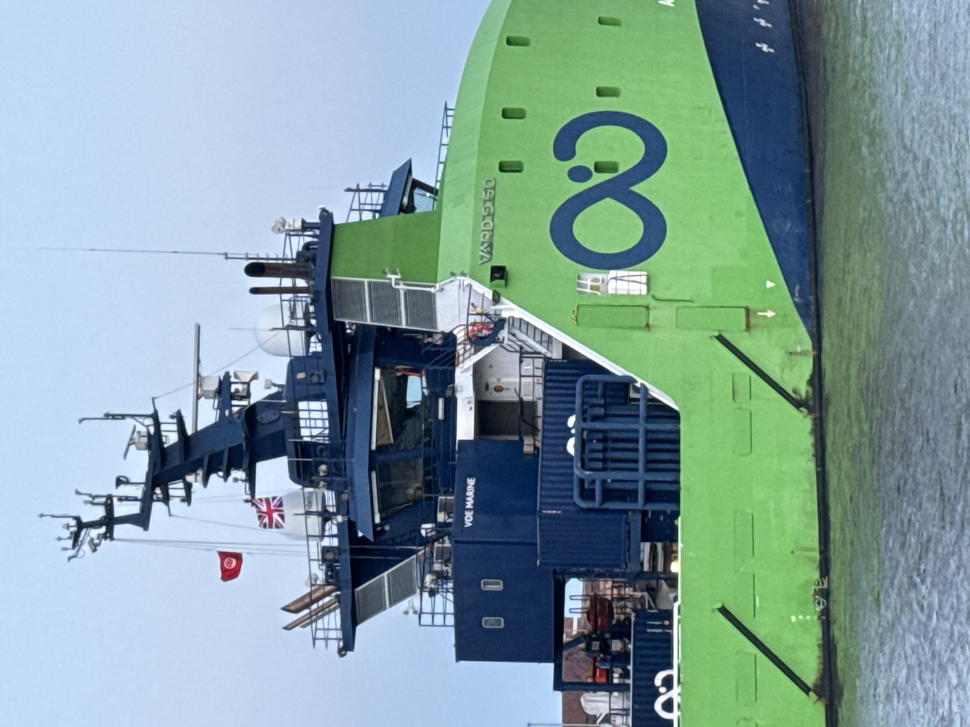 The image shows the Voe Marine ship in Great Yarmouth, United Kingdom. The ship is painted in green and blue. A large number "8" is painted on the green part of the ship. The text "VARD 9150" is written on the green part of the ship near the number 8. The ship has a complex array of antennas and communication equipment on its upper deck. Flags of the United Kingdom and Tunisia are visible on the ship's mast. The ship is docked in the water, with the shoreline visible on the right side of the image.
