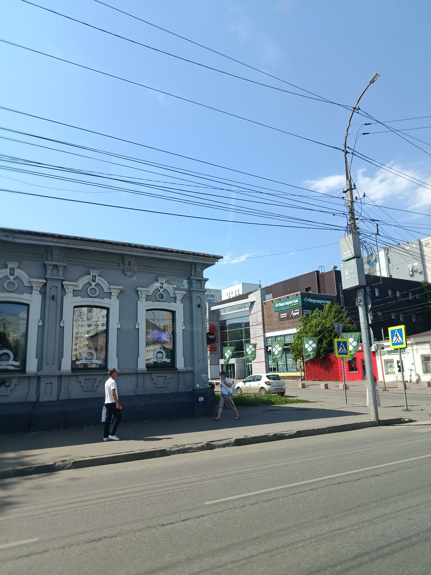In Saratov, Russia, on a clear, sunny day, the image captures a street scene. In the foreground, a man in a white t-shirt and dark trousers walks along the sidewalk. Further down the sidewalk, a woman in a patterned skirt and light-colored top walks with a child. The street is paved, with a gray curb separating it from the sidewalk.
The buildings lining the street vary in architecture. To the left, a gray, two-story building with ornate white trim and arched windows is visible. Inside the windows, reflections of buildings are seen, along with text on the glass indicating "Суши" (Sushi) and "Пицца" (Pizza). To the right, a modern, multi-story building with a glass facade and green banners hangs. The banners feature a white cloverleaf design and the text "Перекресток" (Perekrestok), a supermarket chain. A red wall is also prominent in this section.
Overhead, a complex network of electrical wires stretches across the frame, supported by a tall utility pole adorned with streetlights and various attachments. Two pedestrian crossing signs, featuring a person walking, are placed on the sidewalk. A white car is parked on the grass area in front of the modern building. The sky is a bright blue with a few scattered white clouds.