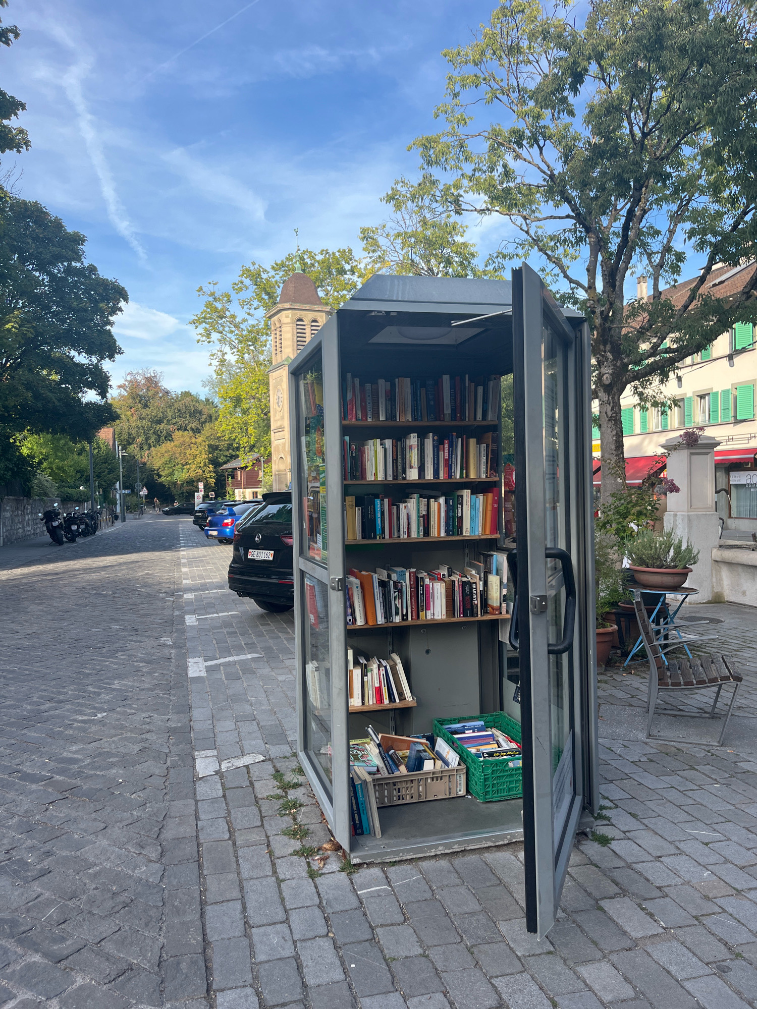 This image captures a street scene in Geneva, Switzerland, featuring a repurposed telephone booth that now serves as a small outdoor library. The booth, made of glass and metal, is open, revealing multiple shelves filled with books. A black SUV with the license plate "GE 801162" is parked in a designated spot on the cobblestone street in front of the library. To the left, a row of parked motorcycles is visible. In the background, a church steeple and building with green shutters suggest a European urban environment. The weather appears to be clear with a blue sky and scattered clouds, and the presence of trees indicates it is likely autumn.