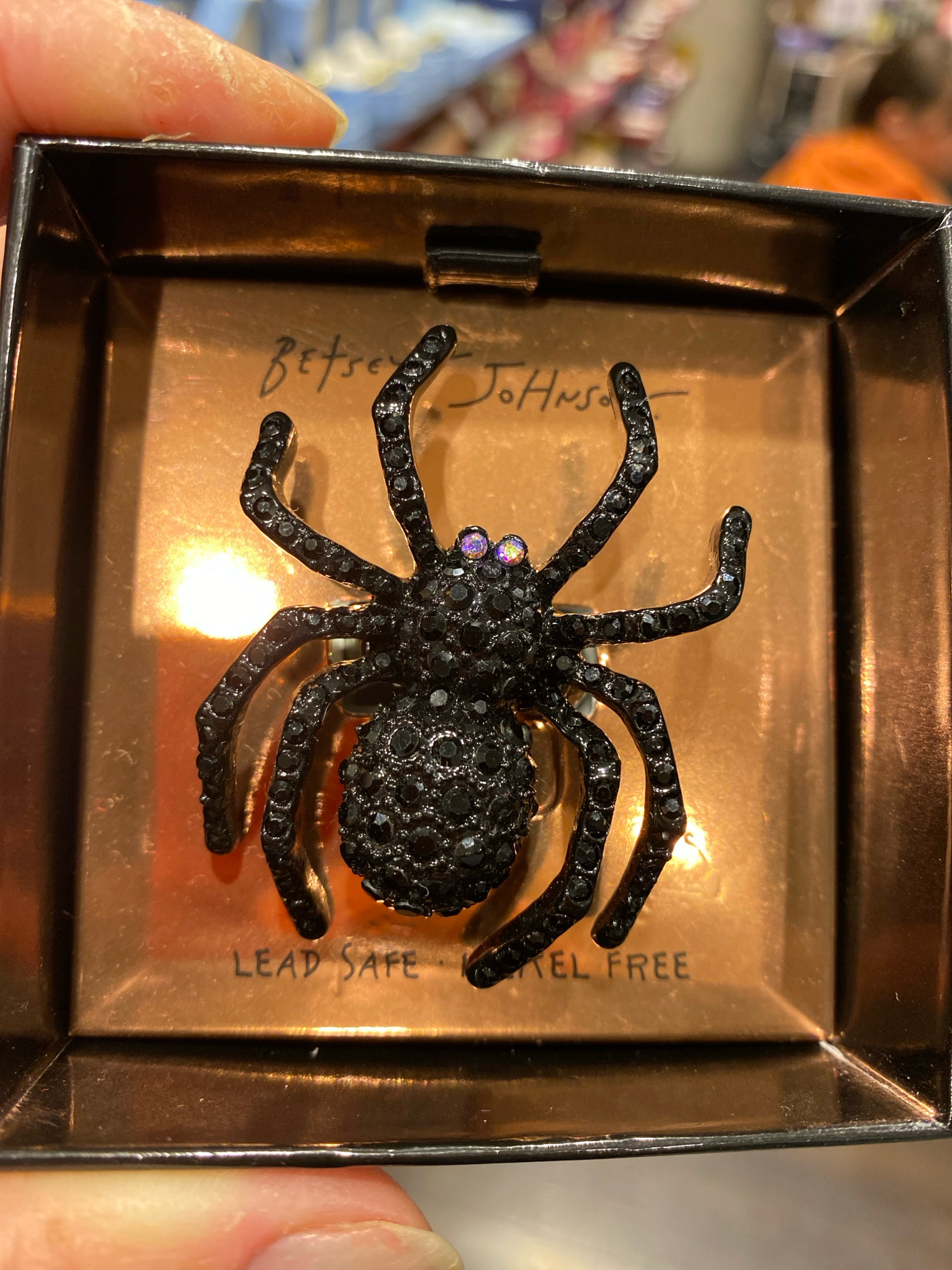 In a square, copper-colored box, a black spider brooch is displayed. The spider is adorned with small, black rhinestones and has two iridescent purple eyes. The inside of the box features handwritten text that reads "Betsy Johnson." Towards the bottom of the box, text printed in black states "LEAD SAFE NICKEL FREE." The box itself is black with a metallic sheen. The image is captured from a slightly overhead angle, with the hand of a person holding the corner of the box visible in the foreground. The background is blurred but suggests an indoor setting with retail displays. The scene does not depict any elements related to Vienna, Austria.