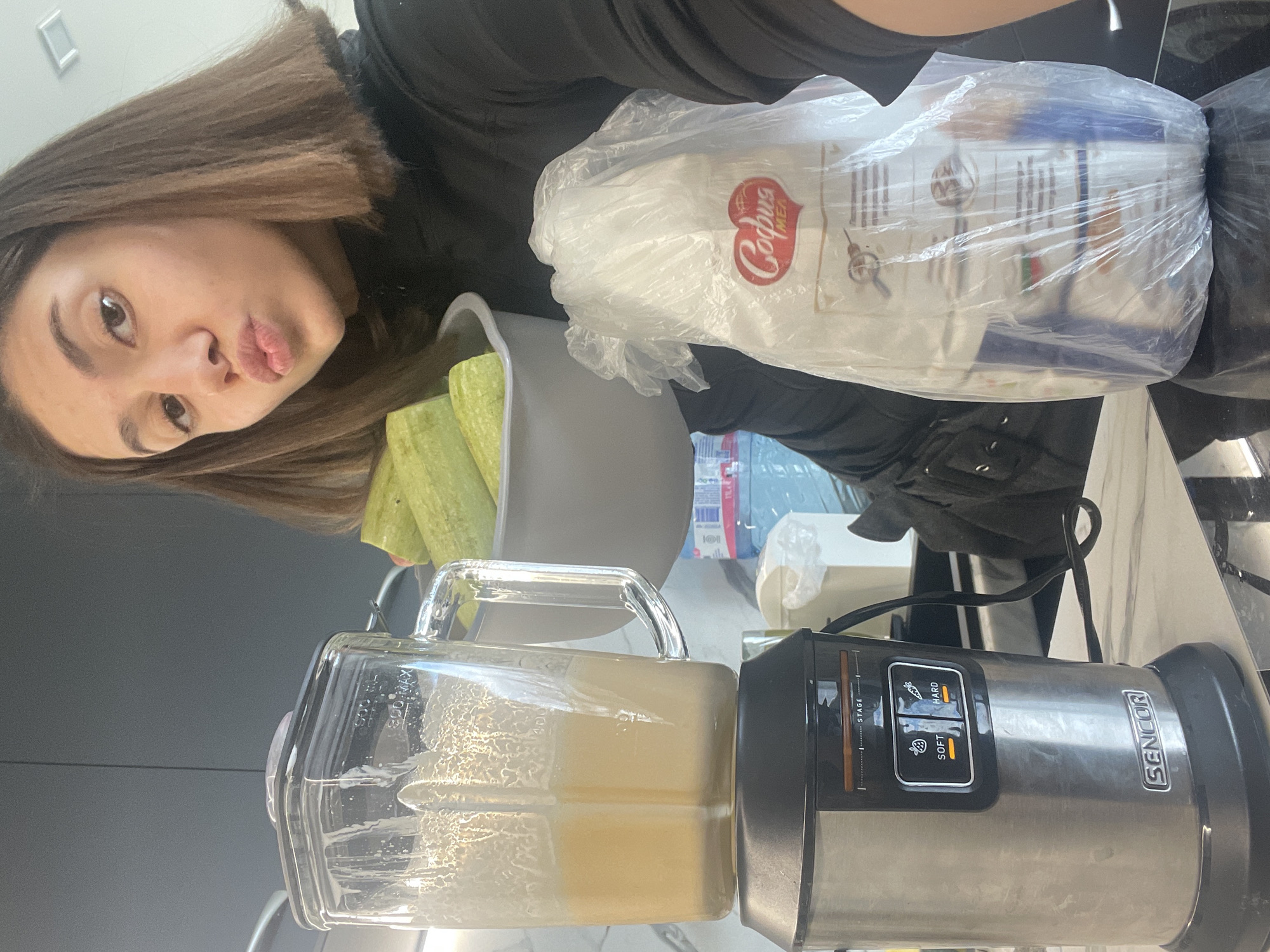 A woman with shoulder-length brown hair and light skin is holding the camera to take a selfie in a kitchen. She is wearing a black top and making a kissy face. In the foreground, there is a Sencor blender filled with a light yellow liquid. Behind the blender, there's a gray container holding zucchini. To the upper right, there is a sealed package of Corpus Sofia brand food, likely from Bulgaria, as Sofia is the capital city. A water bottle is visible behind the food package. The kitchen has gray cabinets and a light-colored countertop.