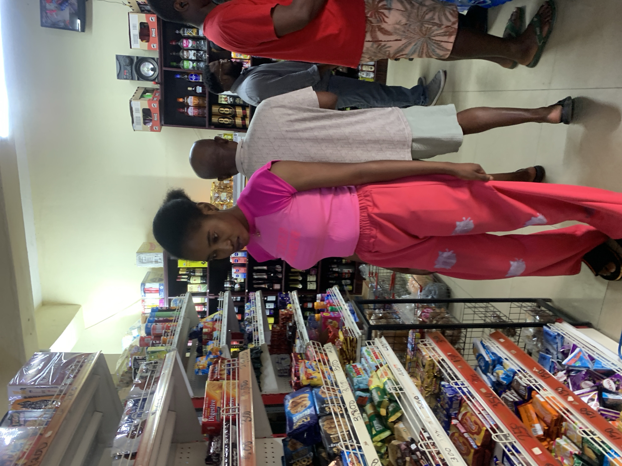 The image depicts the interior of a convenience store, likely located in Abuja, Nigeria. The store features shelves stocked with various food items, beverages, and household goods. A young woman wearing a pink top and pink trousers with white floral patterns stands in the foreground, facing to the right. Behind her, a man in a light-colored patterned shirt and shorts is visible. Further back, another man in a grey shirt and jeans stands to the right of him, and a man in a red t-shirt and patterned shorts is on the far right. The shelves are well-stocked with products, including packaged snacks, drinks, and bottles of alcohol on a higher shelf. The lighting suggests an indoor environment, possibly with fluorescent lights overhead. The overall scene suggests a retail environment where customers are browsing or waiting.