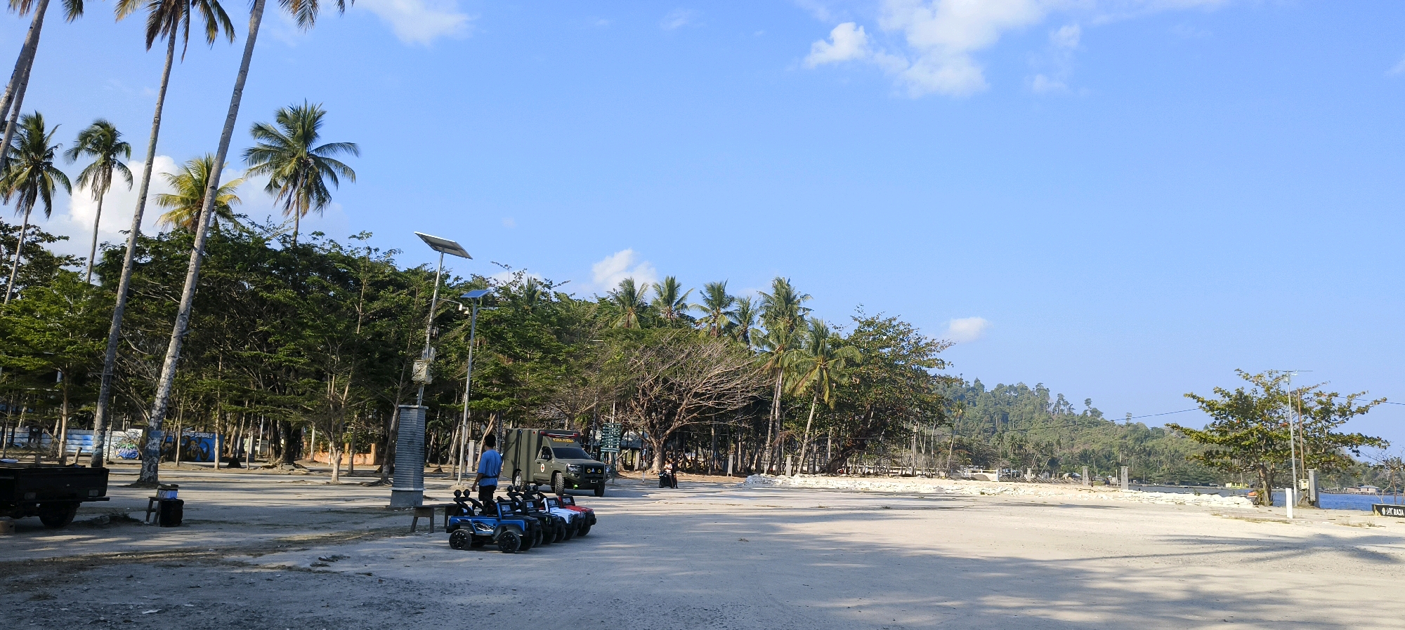 The image depicts a bright, sunny day at a beachside area in Waisai, Indonesia, likely a parking or recreational zone. The foreground is a sandy and gravelly expanse with distinct shadows cast from palm trees lining the left side of the frame. Several small, ride-on toy cars, primarily in blue and red, are lined up. To the right of these toy cars, a man in a blue shirt stands near a dark green, military-style truck. Further back, a dense line of tropical trees and palm trees forms a backdrop, with some appearing more sparse and dry than others. Beyond the trees, a green, hilly landscape is visible, leading to the blue ocean. The sky is clear and bright blue with a few wispy clouds. On the left side of the image, a dark trailer is parked. There are also solar-powered streetlights visible. The overall impression is of a tropical coastal location, possibly a tourist spot or a public beach area.