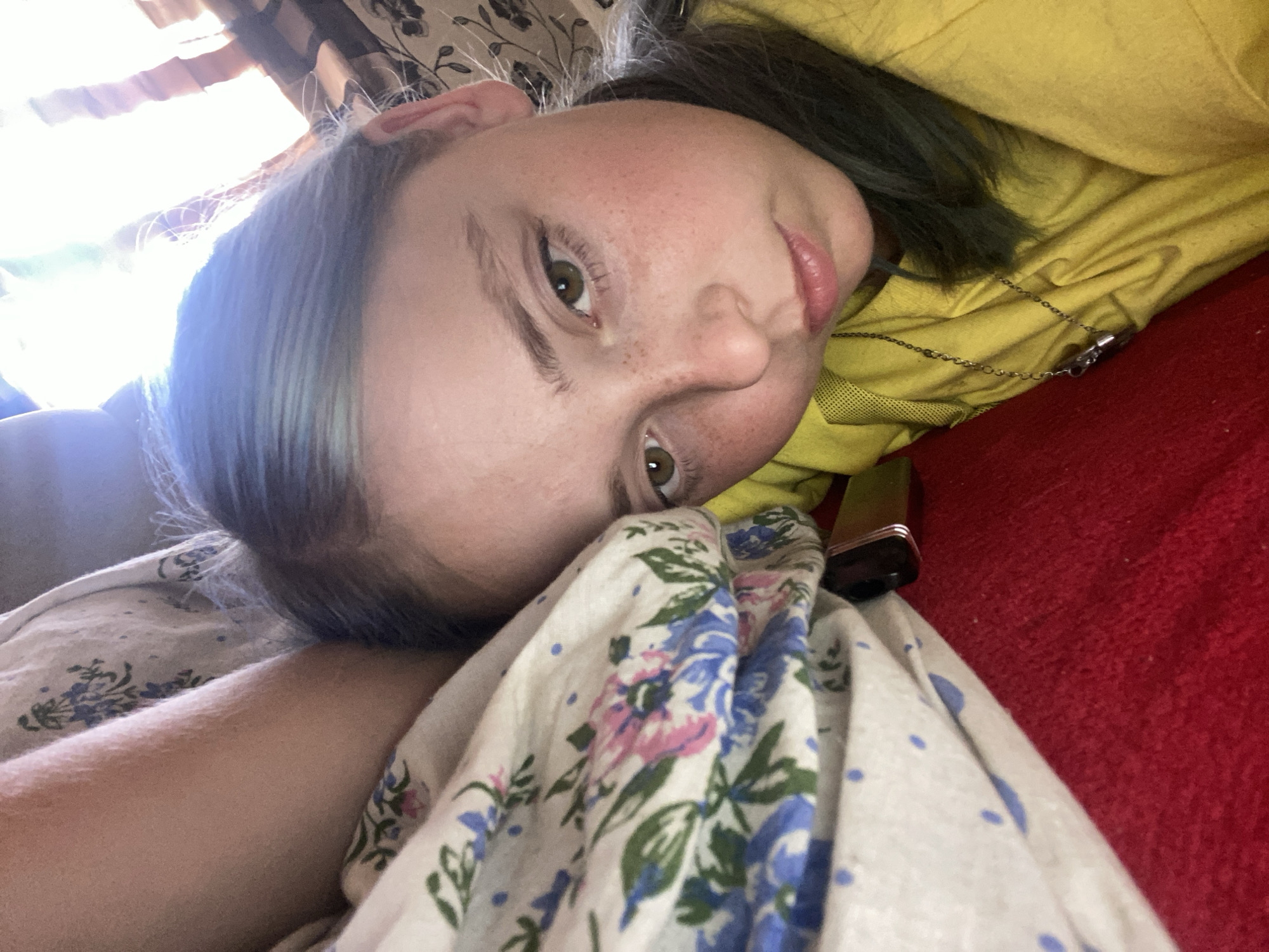 The image shows a young person lying on their side, with their head resting on a floral-patterned blanket. They are wearing a yellow garment and have dark hair with streaks of blue or green. Their eyes are open and looking towards the camera, and they have freckles on their cheeks. The lighting suggests it is daytime, with sunlight streaming in from the left. The background appears to be indoors, with hints of curtains and a red surface that could be a couch or bed. The overall mood is relaxed and informal. There are no visible text elements in the image. The context provided states the location as Nova Praha, Ukraine.
