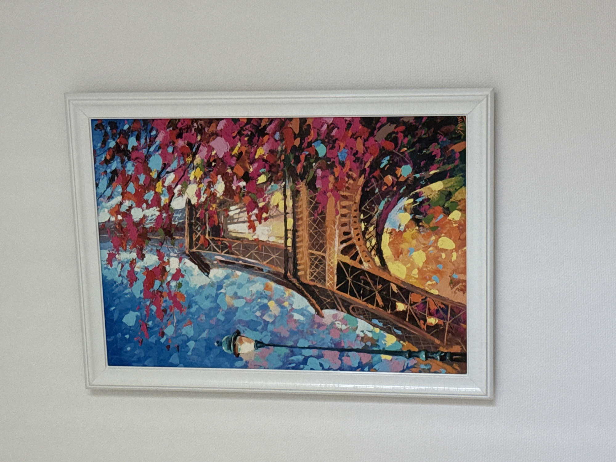 The image shows a framed painting of the Eiffel Tower hanging on a textured, white wall. The painting is in a vibrant, impressionistic style with thick brushstrokes of color. The Eiffel Tower is depicted in shades of brown and orange, with the surrounding sky in bright blue and pink. There is a streetlamp in the foreground, also painted in a similar colorful style. The signature "Spiro" is visible in the bottom right corner of the painting. The context of the city of Burgas, Bulgaria is not present in the image itself, and therefore cannot be described.