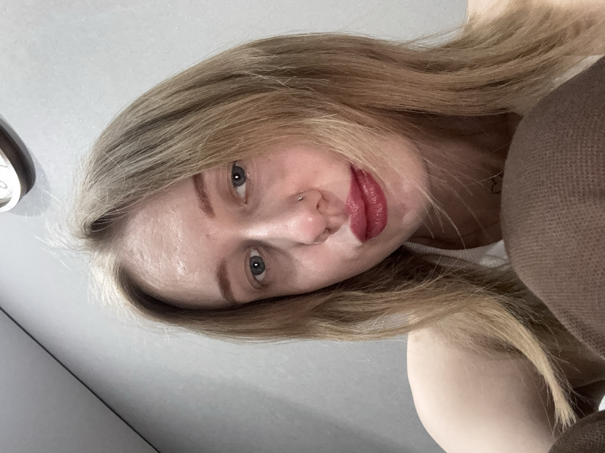 An indoor portrait captures a female subject looking directly at the camera. The background consists of a plain light gray wall, with a partial view of a wall-mounted clock featuring a silver rim in the upper right. The subject has long, light blonde hair. Her left eye (viewer's right) is blue and fully visible, while her right eye is partially obscured by hair. Her lips are coated with dark red lipstick and are slightly puckered. A silver stud piercing is present in her left nostril. A small, dark heart-shaped tattoo is visible on her upper chest, slightly to the left. She is wearing a white or light-colored strap top, and a brown, textured fabric is visible in the lower frame, indicating another garment or object. The scene is located in the city of Novoaleksandrovka, Russia.