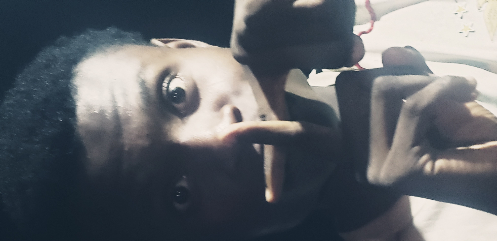 A close-up, dimly lit portrait shows a young Black person with dark, curly hair and dark eyes looking directly at the camera. Their right index finger is held to their lips, and their left hand is raised, with fingers splayed. The person is wearing a white garment, possibly a t-shirt. The background is dark, suggesting it is nighttime or a poorly lit indoor environment in Narayi, Nigeria. The image is blurry, with some motion blur evident, particularly in the hands.