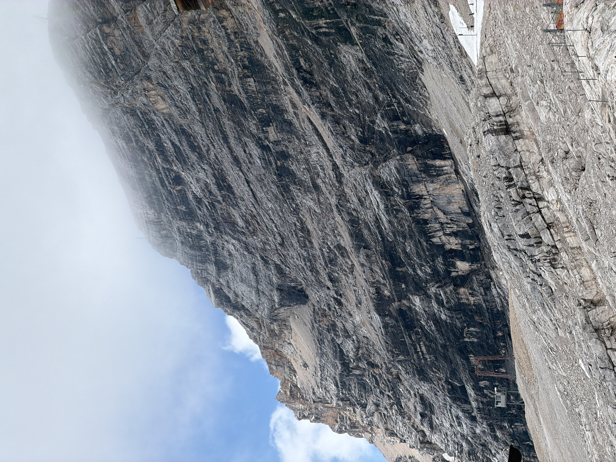 The image shows a steep, rocky mountain face, likely part of the Zugspitze massif, near Garmisch-Partenkirchen, Germany. The mountain is characterized by layered rock formations, with dark streaks and patches of lighter rock. The upper part of the mountain is obscured by thick, white clouds, with patches of blue sky and white clouds visible on the left side. In the foreground, the terrain is also rocky and uneven, with some sparse vegetation. A section of a ski slope with a rope barrier is visible on the right, and a few small figures, possibly skiers or hikers, are present on the snow. Lower down on the mountain, there is a small, grey building with a red metal structure, possibly part of a cable car station or maintenance facility. The overall impression is of a rugged and imposing alpine landscape under a cloudy sky.