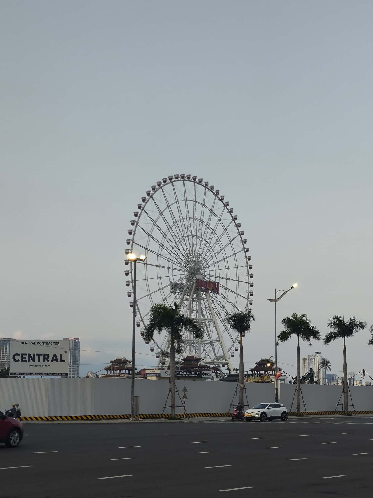 The image depicts an urban scene in Đà Nẵng, Vietnam, specifically facing the Asia Park area. In the center, a large white Ferris wheel, identified as the "SUN WHEEL" by text on its hub, dominates the middle ground. Its capsules are red. Several tall palm trees are situated in front of the Ferris wheel. Behind a white construction barrier, traditional Vietnamese-style pagoda roofs and other park structures are visible.
To the left, a large billboard for "GENERAL CONTRACTOR CENTRAL" (www.centralcons.vn) is prominent. Further left and in the background, modern high-rise buildings are visible. The foreground consists of a multi-lane asphalt road with white lane markings. A yellow and black striped barrier separates the road from the construction area. Streetlights with illuminated lamps are present on both sides of the road, particularly one on the left in front of the Ferris wheel and another on the right.
Vehicles observed include a partial view of a red SUV on the far left, a white SUV/crossover stationary or moving slowly in the middle lane, and a red scooter positioned next to the white SUV, operated by an individual wearing a light-colored helmet. The sky is uniformly light grey, indicating overcast conditions or twilight hours, with artificial lights already active.
