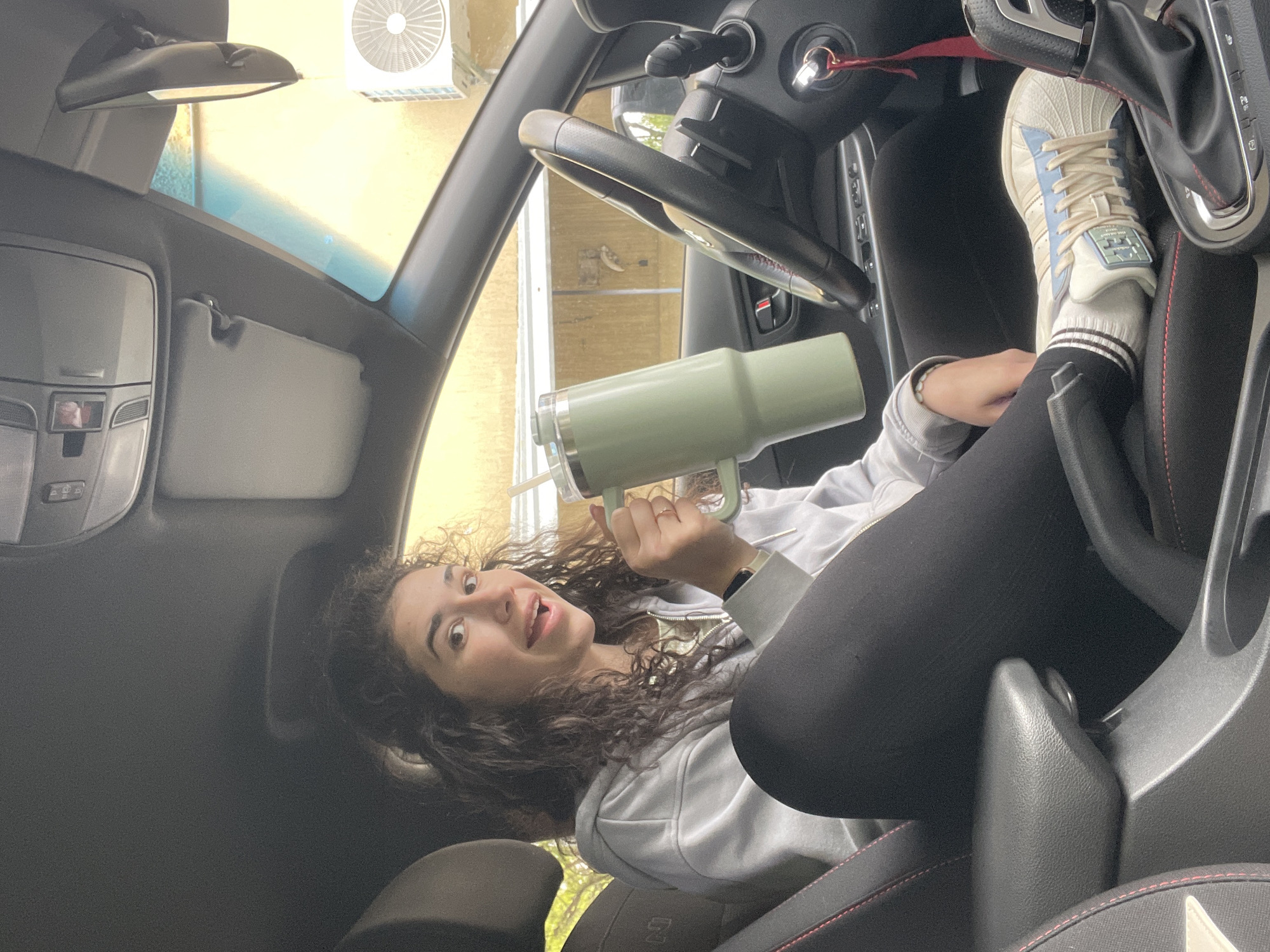 A young woman with dark, curly hair and a playful expression is photographed inside a car, appearing to be lying on her side due to the image's vertical orientation. She looks directly at the camera with a slight smile and her tongue slightly out, holding a large, light green thermal mug with a handle in her right hand. She is wearing a light grey hooded sweatshirt and black leggings. Her left foot, adorned with a white Adidas Superstar sneaker featuring light blue accents and beige laces, and white socks with black stripes, is propped up near the car's gear shifter. The visible text "Adidas" and its logo are clearly seen on the shoe's tongue.
The car's interior includes a steering wheel, keys in the ignition, and black seats with red stitching, one of which displays an "N" logo. Bright daylight illuminates the scene, suggesting it is daytime. Outside the car windows, an air conditioning unit is visible on a light-colored building, indicating the car is parked outdoors. The overall mood of the image is casual and cheerful. This scene takes place in Sofia, Bulgaria.