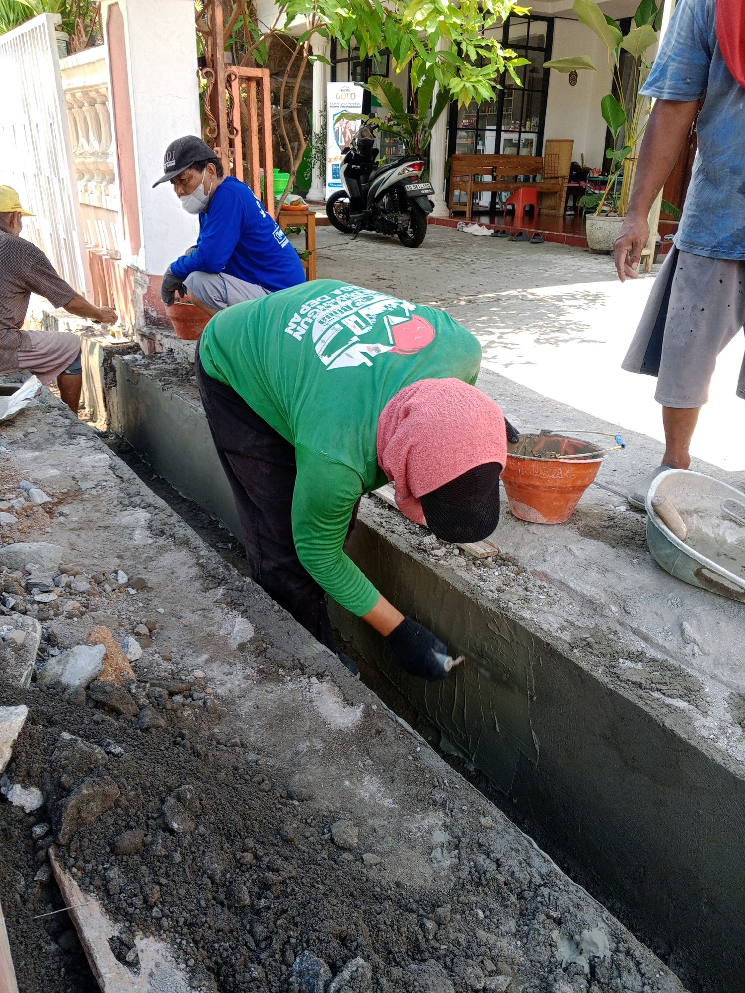 In Giritirto, Indonesia, three construction workers are engaged in a drainage ditch repair. Two individuals are actively working, one in a green long-sleeved shirt and black pants, applying cement to the side of the ditch with a trowel, and another in a blue long-sleeved shirt, light pants, and a black baseball cap, crouching and appearing to supervise or prepare materials. A third worker, wearing a brown shirt and pink shorts, is partially visible in the background, also appearing to be involved in the construction. The environment is outdoors, with a paved ground surface, exposed concrete structures, piles of dirt and rocks, and vegetation visible. A motorcycle is parked in the background, and a building with a white facade is present. The scene suggests a typical day of manual labor on a construction site.