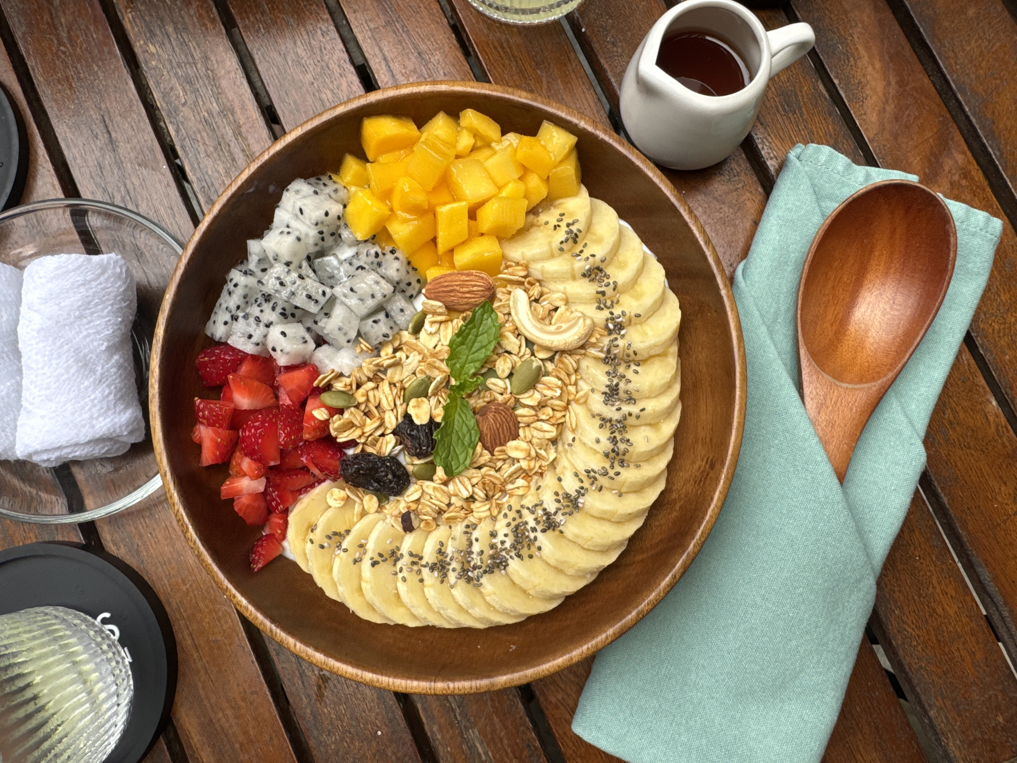 The image displays a beautifully arranged breakfast or brunch bowl set on a warm, slatted wooden table, indicative of a natural and inviting dining environment, possibly in Đà Nẵng, Vietnam.
The central subject is a large, rustic wooden bowl filled with a colorful and healthy assortment of food. The bowl's contents are divided into sections: vibrant yellow diced mangoes occupy one quadrant, followed by white diced dragon fruit with its characteristic black seeds. Another section features bright red chopped strawberries, while thinly sliced bananas, generously sprinkled with black chia seeds, form a semi-circular border around the remaining contents. At the heart of the bowl, a mound of golden granola is visible, adorned with various nuts and seeds, including an almond, a cashew, pumpkin seeds, and dark raisins, all garnished with a fresh green mint leaf.
Around the main food bowl, several complementary items are present. To the upper right, a small white ceramic pitcher contains a dark liquid, likely syrup or honey. Adjacent to it, a large, smooth wooden spoon rests gracefully on a neatly folded light blue linen napkin. To the left, a clear glass bowl holds two clean, white folded hand towels, suggesting a refreshing amenity. Part of a textured glass, possibly containing a beverage like water or juice, is visible on a black coaster in the bottom left corner.
The setting appears to be outdoors or in a brightly lit indoor space during the daytime, given the natural light. The overall presentation suggests a focus on fresh ingredients and a pleasant, relaxed dining experience. No people are visible, and there is no readable text on any of the objects.
