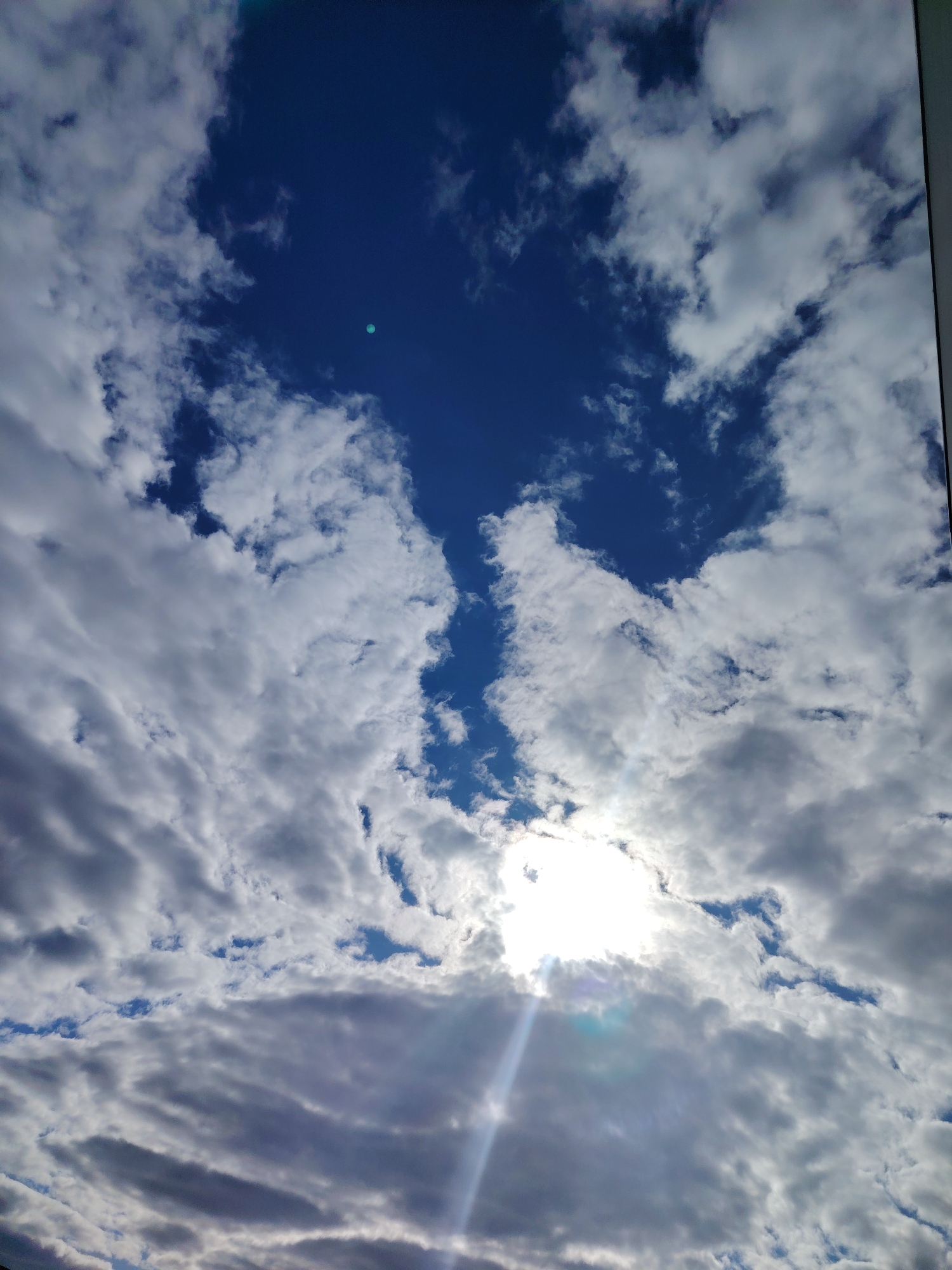 The image shows a sky with clouds and patches of blue sky visible. The sun is shining through the clouds in the lower center of the frame. The clouds vary in thickness and coloration, ranging from bright white to gray. There is no indication of the location being in the city of Noviy, Russia, or any signs of people, subjects, objects, or actions.
