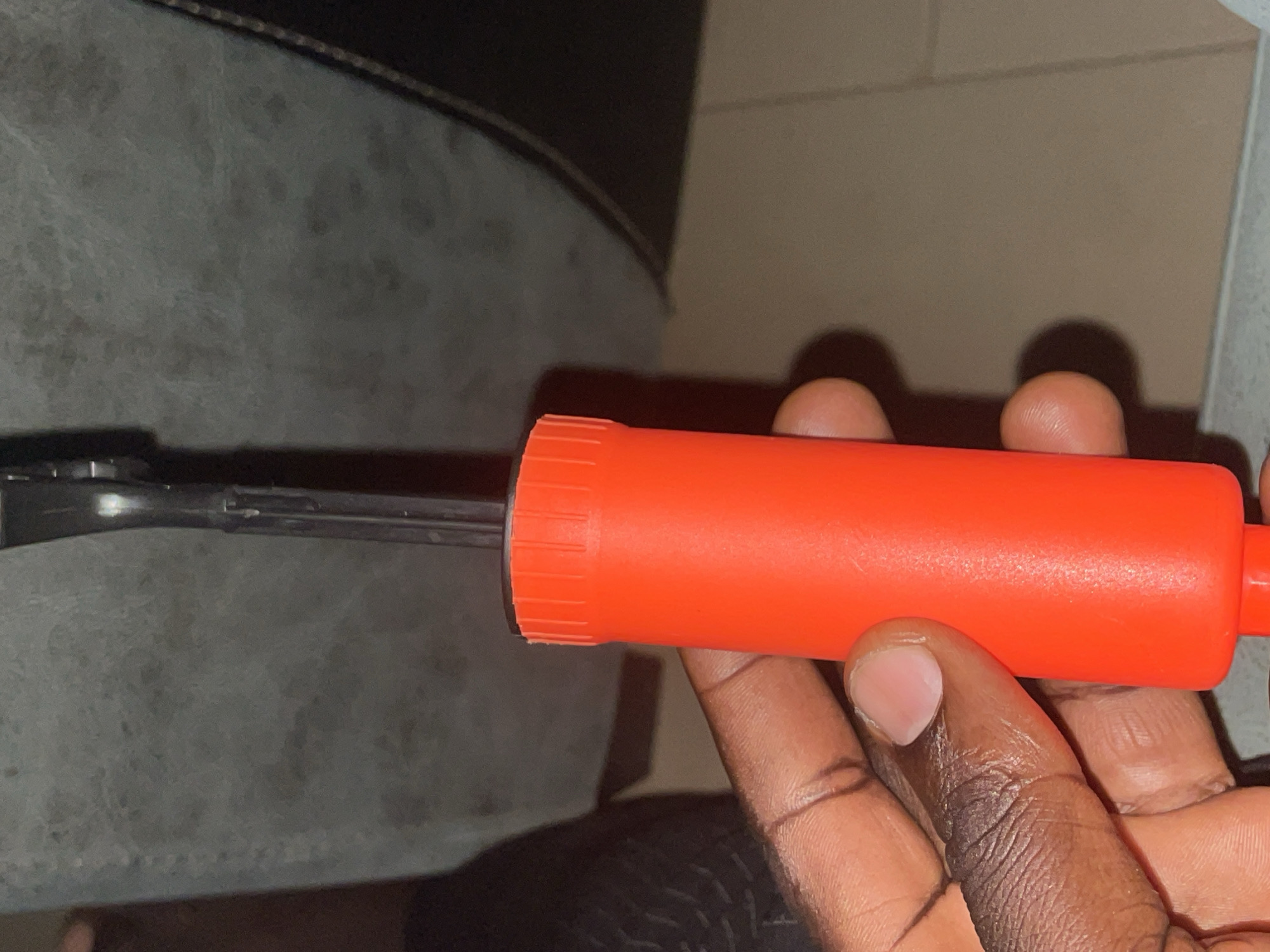 A close-up shot shows a person's hand holding an orange ball pump. The pump has a ribbed top and a smooth cylindrical body. The hand is dark-skinned, with visible skin texture and fingernails. The background is out of focus, appearing to be a textured gray surface, possibly a wall or fabric, and a tiled floor. The lighting suggests an indoor environment. The scene is not specific to any location in the city of Jos, Nigeria, as no identifying landmarks or context are present.
