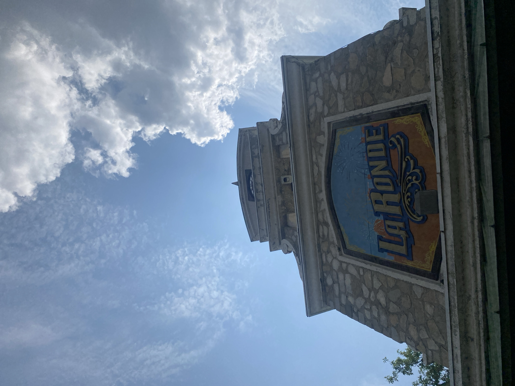 The image is a low-angle shot of the La Ronde amusement park entrance tower, located on Île Sainte-Hélène in Montreal, Canada. The tower is constructed of stone with decorative architectural elements and features a large, colorful sign displaying "LA RONDE" with fireworks graphics. The sky above is bright blue with scattered white clouds. Some greenery from trees is visible on the left side of the frame. The photo is taken from a ground-level perspective looking upwards towards the tower.