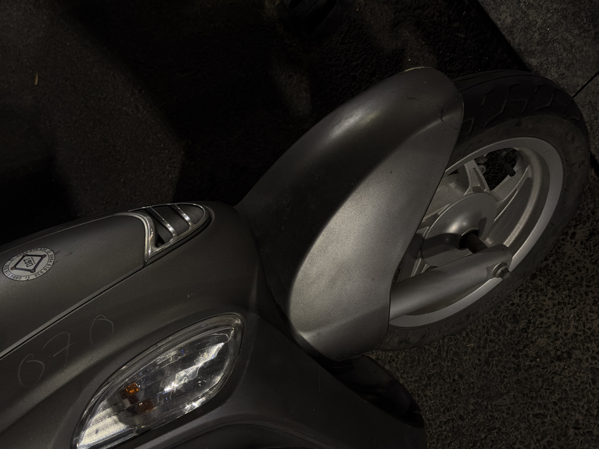 This media file shows a close-up, high-angle shot of a silver scooter's front portion, focusing on the fender, headlight, and part of the wheel. The scooter is parked on an asphalt surface, possibly a street or parking lot, with some dark, textured ground visible in the background. The lighting suggests it might be nighttime or a dimly lit area, with a strong light source creating reflections on the scooter's metallic surfaces.
The scooter itself appears to be a typical model, with a sleek, modern design. The front fender curves over the wheel, and the headlight unit is prominent, featuring an orange bulb inside a clear lens. There are some white chalk markings on the scooter's body, appearing to be numbers or symbols, though they are not entirely clear. One marking, "370," is somewhat legible.
On the scooter's body, near the front, there is a round sticker or emblem. It has a diamond shape in the center with text around it. The visible text on the sticker includes "DET" in the center, and partially visible numbers and letters around the edge such as "0984 619 836".
The scene is static, with no people or activity evident. The focus is purely on the details of the scooter and its immediate surroundings. The lack of context clues like street signs or specific architecture makes pinpointing an exact location within Da Nang difficult, but the overall impression is of an urban setting. The quality of the image, with its strong contrast and shadows, suggests it may have been taken with a mobile phone camera.