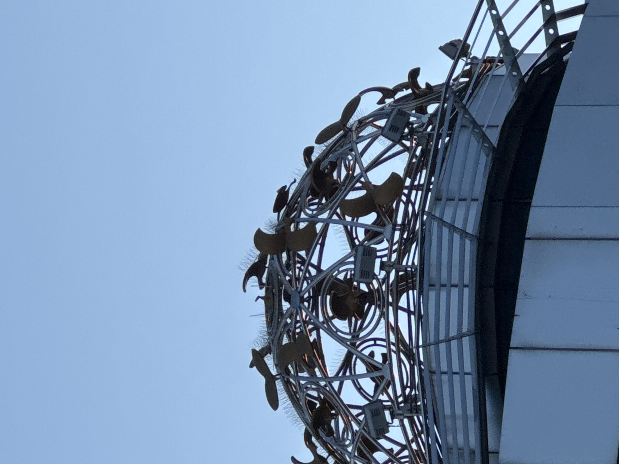 The image shows the sculptural installation "Compass of Rose", located in Burgas, Bulgaria. The sculpture features metal birds and circles arranged within a spherical, lattice-like metal frame. This structure is attached to a tall, modern building with vertical lines. The background is a clear, light blue sky.