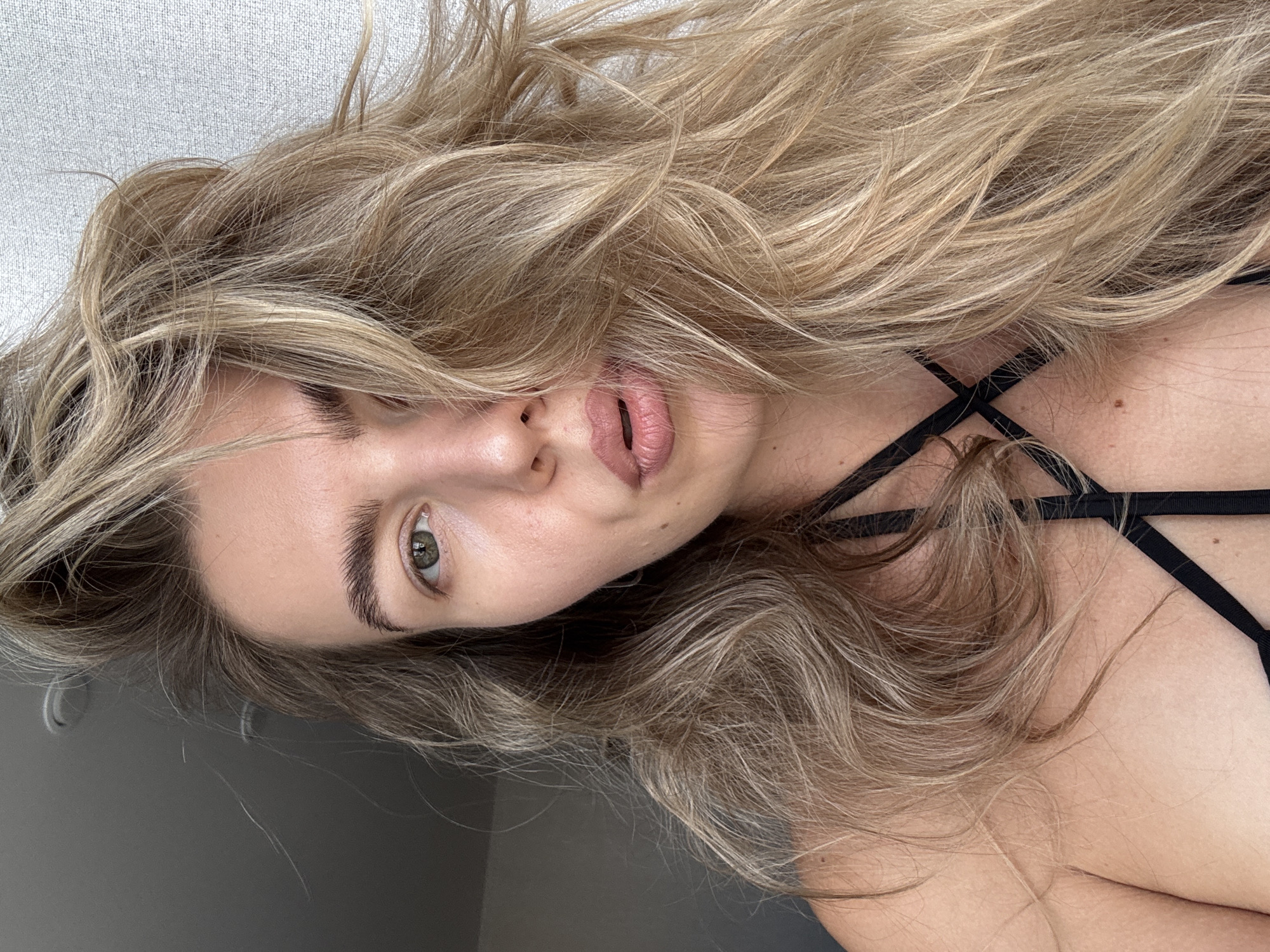 A close-up, medium shot captures a woman's face and upper body against a textured gray background. Her blonde hair, with streaks of lighter blonde, is tousled and partially obscures her face, falling across her right eye and mouth. She is looking directly at the camera with a neutral expression. Her makeup is minimal, with emphasis on her eyebrows and lips, which are slightly parted and appear to have a light pink lipstick. She is wearing a black halter top with thin straps that cross at the chest. The lighting is soft, creating subtle shadows and highlights on her skin. There are no discernible background elements that indicate a specific city or location.