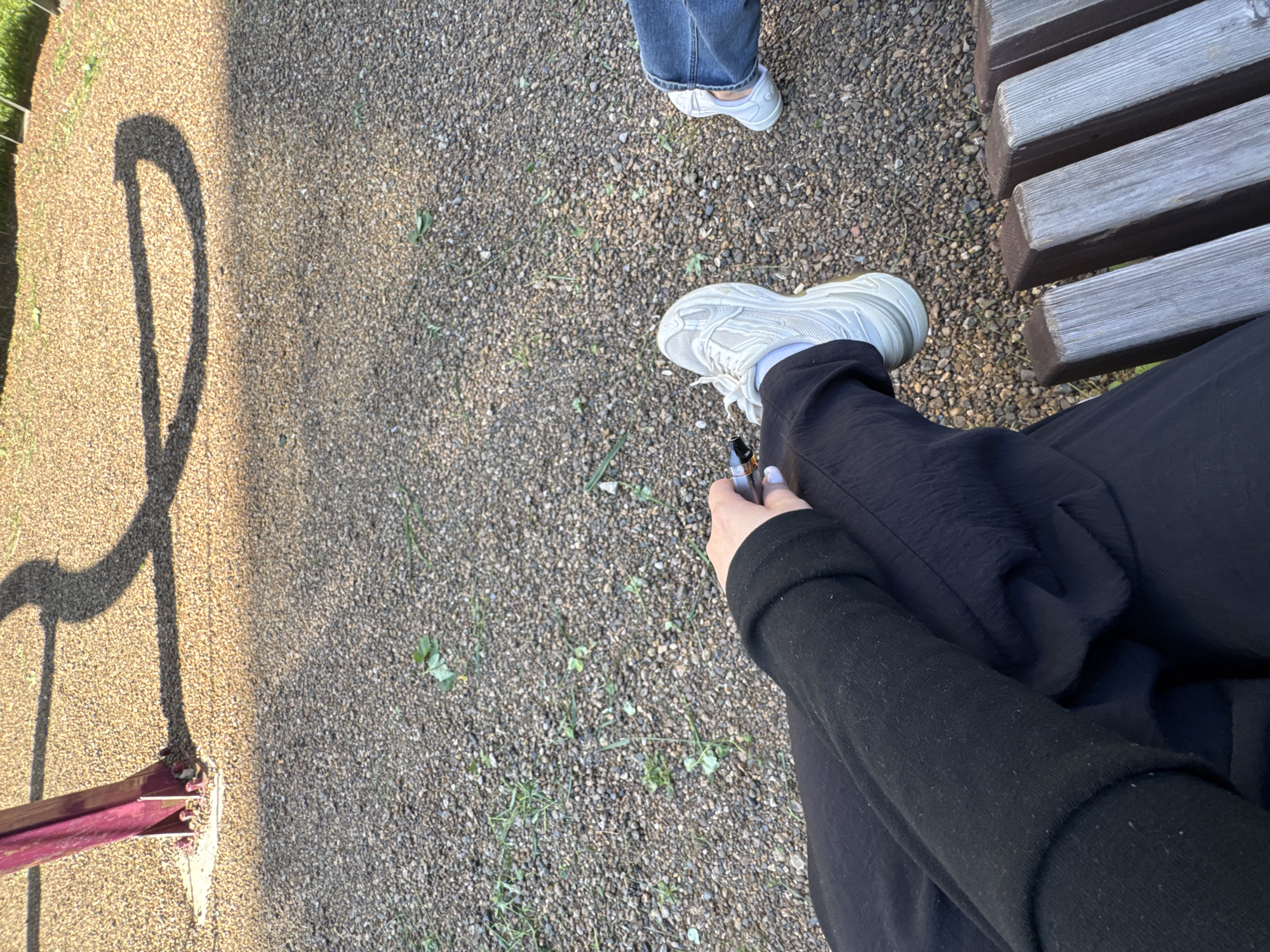 A low-angle, close-up shot captures two individuals sitting on a wooden bench in what appears to be a park or playground area. The ground is covered with small, gray pebbles and sparse patches of green grass. In the upper left quadrant of the image, a portion of a burgundy-colored playground structure is visible, casting a curved shadow onto the pebbled ground.
One individual, positioned on the left side of the frame, is wearing black pants and a black long-sleeved garment. They are holding a small, metallic object, possibly a vape pen, in their right hand, with their thumb and index finger extended. Their left foot is clad in a white sneaker with a chunky sole.
The other individual, on the right side of the frame, is wearing blue jeans and a white sneaker, similar in style to the other. Their legs are partially visible.
The lighting suggests it is daytime, with the sun casting distinct shadows from the playground structure. The scene is devoid of any identifiable landmarks or specific elements that would pinpoint the location to the city of Perm, Russia.