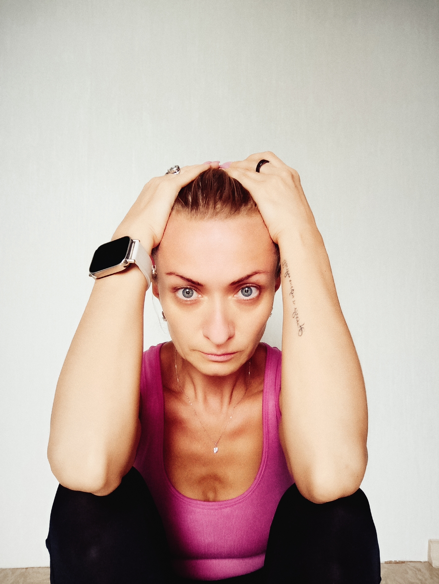 A woman with blonde hair pulled back is sitting with her hands on her head. She is wearing a pink tank top and black pants. On her left wrist is a smartwatch with a white band. On her right arm, there is a tattoo on her forearm. She has blue eyes and a serious expression. The background is a plain white wall. The scene is indoors. This is not in Moscow, Russia.