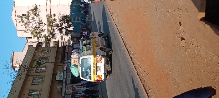 A white and yellow mini-van, likely a Matatu, sits on a city street, its roof laden with luggage. Buildings of varying heights line the left side, one adorned with a partially bare tree. A busy scene unfolds in the background, suggesting a market or gathering place. The foreground is dominated by a dusty, worn sidewalk or roadside. The lighting suggests it's a bright, sunny day.