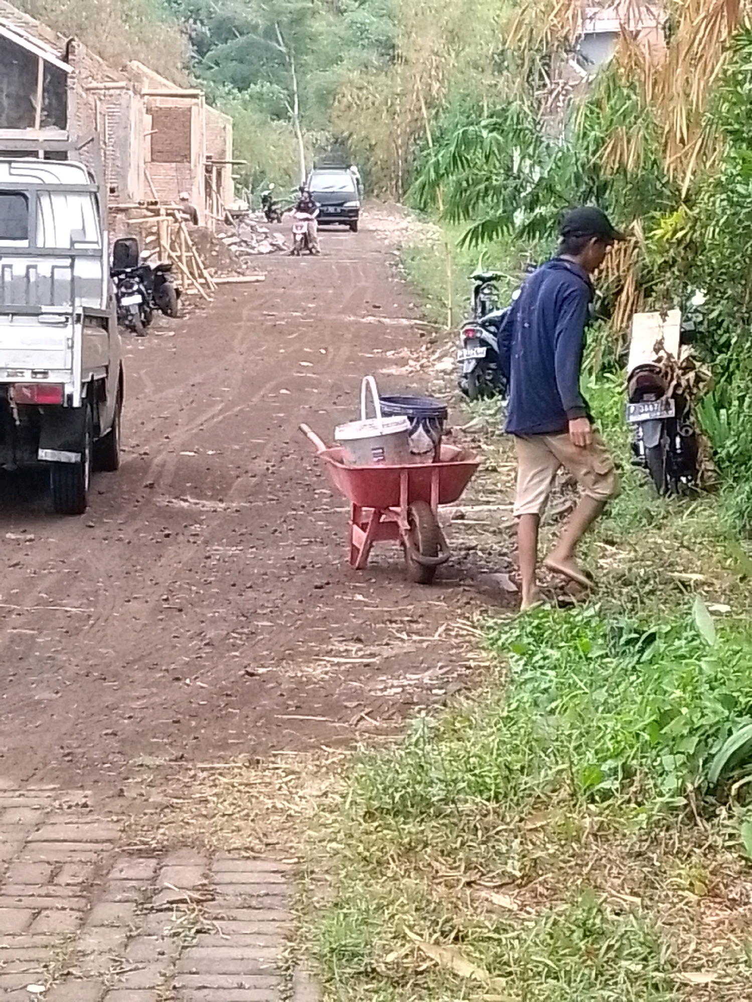 The image depicts a construction site in Jember, Indonesia, likely a residential area given the presence of building materials and unfinished structures. A light-colored utility truck is parked on the left side of a dirt road, which appears to be under construction or in a state of disrepair, indicated by the uneven surface and tire tracks. A red wheelbarrow filled with two buckets, one white and one dark, is positioned in the middle of the road. A person wearing a dark blue long-sleeved shirt, tan shorts, a black baseball cap, and no shoes is walking away from the wheelbarrow towards the right side of the frame, which is overgrown with lush green vegetation, including bamboo. Several motorcycles are parked on the left side of the road, near a partially constructed brick building with scaffolding. In the background, a black SUV is visible on the road, and more vegetation covers the surrounding hills. The overall scene suggests ongoing construction work in a semi-rural or developing urban environment.