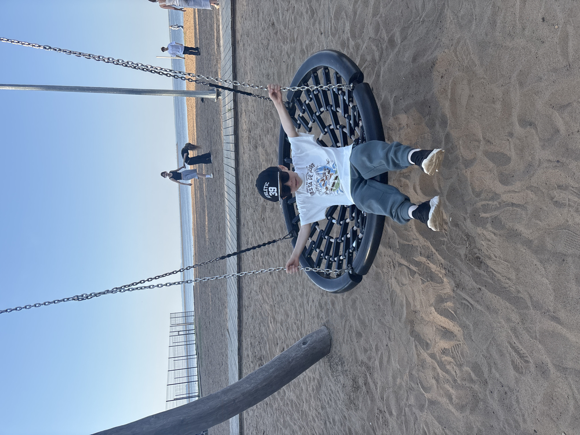 A child is on a circular swing at a beach in Chaikovsky, Russia. The child is wearing a baseball cap, sunglasses, a t-shirt with a graphic, grey sweatpants, and sneakers with black socks. The swing is suspended by chains from a metal structure. There are people walking along the beach and the shoreline is visible in the background. The beach has a wooden walkway and metal fencing. The sky is blue. The location is sandy.