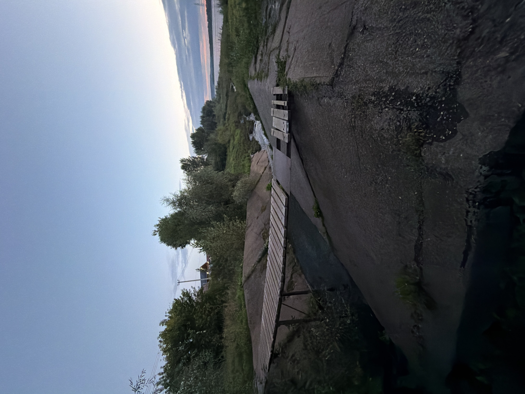 The image depicts a concrete embankment with a small wooden footbridge crossing a narrow channel of water. To the left, a longer wooden bridge is partially visible. Lush green vegetation covers the banks of the channel and the surrounding area. In the background, trees and a few buildings are visible, along with a power line. The sky above is a pale blue with some wispy clouds, suggesting either early morning or late evening. The scene appears to be located along the Kama River embankment in Chaikovsky, Russia, given the characteristic concrete structures and the presence of a large body of water. No people are visible in the frame.