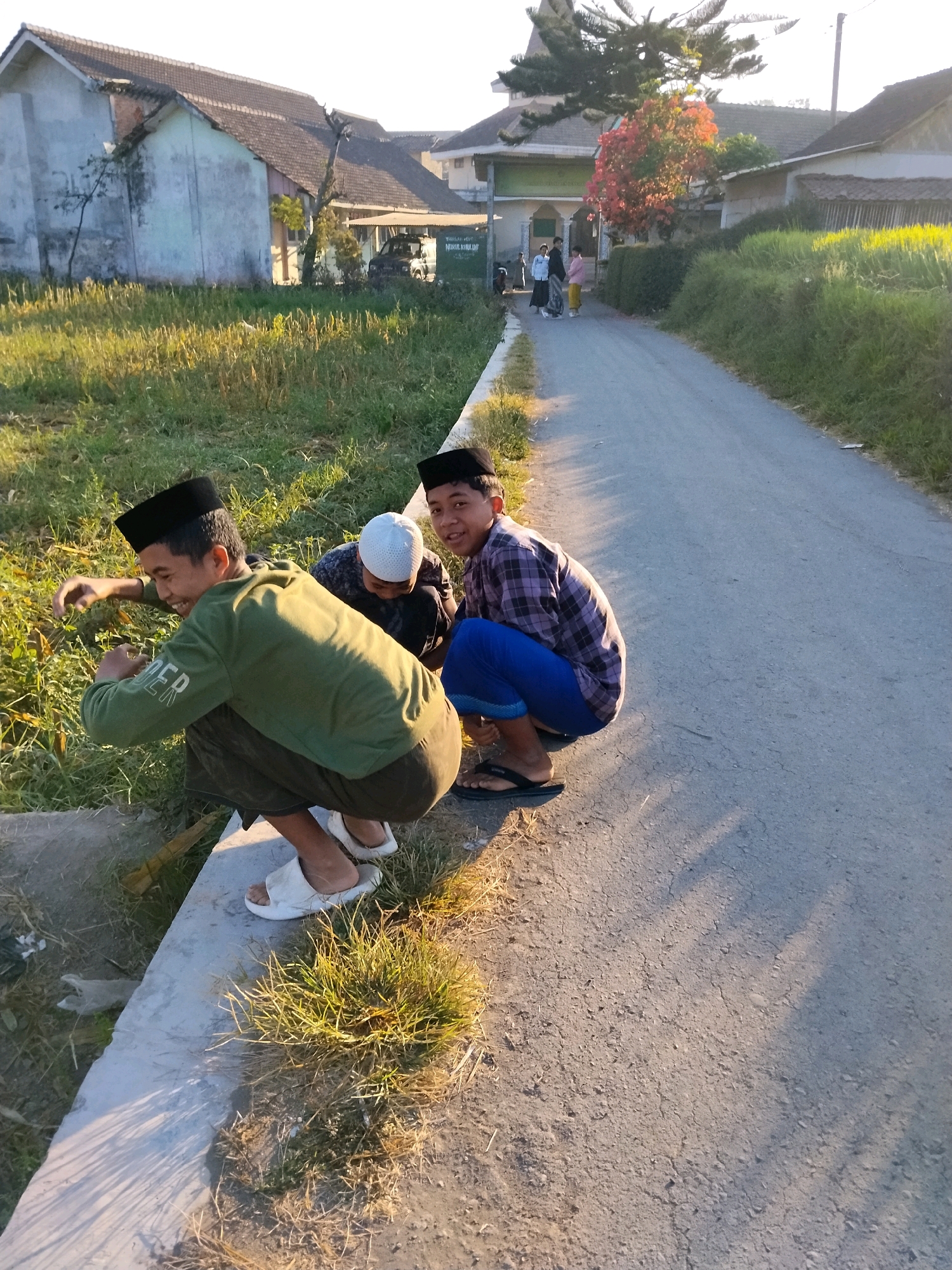 Three young males are crouching outdoors on a sunny day in Tlogosari, Indonesia. They are positioned on a raised concrete embankment next to a paved road. The male on the left, wearing a green long-sleeved shirt and dark pants, is looking down and to his left. In the middle, another male wearing a white skullcap and a patterned shirt is looking down. The male on the right, wearing a plaid shirt and blue pants, is looking at the camera and smiling. A fourth individual, partially visible, is also crouching behind the male on the right. The environment consists of dry grass and vegetation on the embankment and a road with shadows cast across it. In the background, houses with tiled roofs and trees are visible. The overall lighting suggests it is either early morning or late afternoon.
