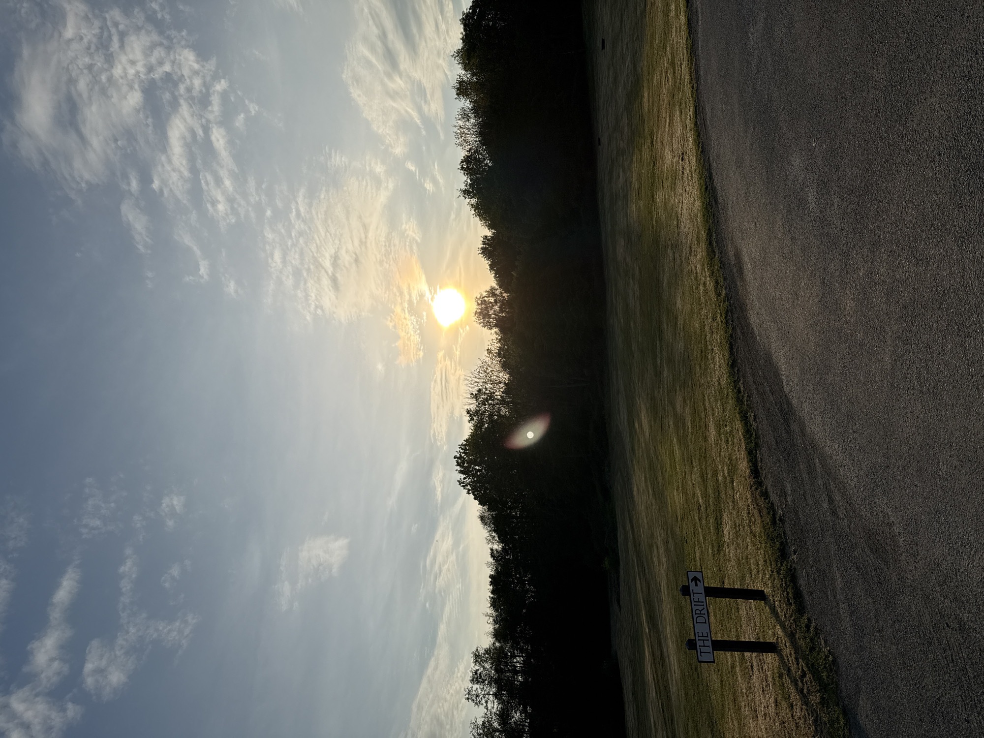 The image shows a landscape at sunset or sunrise. A sign reads "THE DRIFT" with an arrow pointing to the right. This suggests the location is likely near The Drift nature reserve. A road runs along the right side of the image, adjacent to a grassy area. A line of trees is silhouetted against the sun, which is obscured by clouds in the sky. The sky is light blue with scattered white clouds. The overall scene appears peaceful. Based on the sign and knowledge, the location is likely in or near East Suffolk, United Kingdom, where The Drift nature reserve is located.