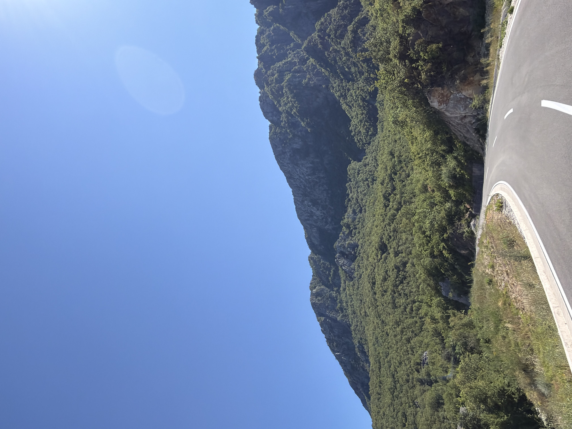The image shows a winding paved road in a mountainous, vegetated area. The road has white dashed lines and a white border on the right side. The left side of the road has dry grass and some bushes. The background is dominated by a large, tree-covered mountain slope with rocky outcrops. The sky is clear and blue, with a bright sun creating a lens flare in the upper right quadrant. The scene appears to be in a rural, natural setting, likely in the vicinity of Bukovik, Montenegro. There are no people or animals visible in the image.