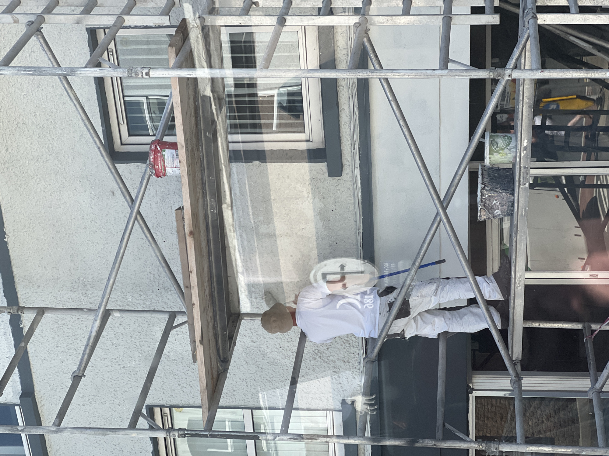 This image captures a person working on scaffolding outside a building in the city of Donegal, Ireland. The individual, dressed in white overalls and a light-colored hat, is actively painting a textured exterior wall. They are holding a large, circular tool, likely for applying or smoothing paint. Scaffolding structures, made of metal poles and wooden planks, surround the worker, indicating ongoing construction or renovation. Various paint buckets and tools are visible on the scaffolding platforms. The building itself has multiple windows, some of which appear to be reflective. The overall scene depicts a typical day of exterior building maintenance or improvement in a presumably urban or semi-urban setting within Donegal.