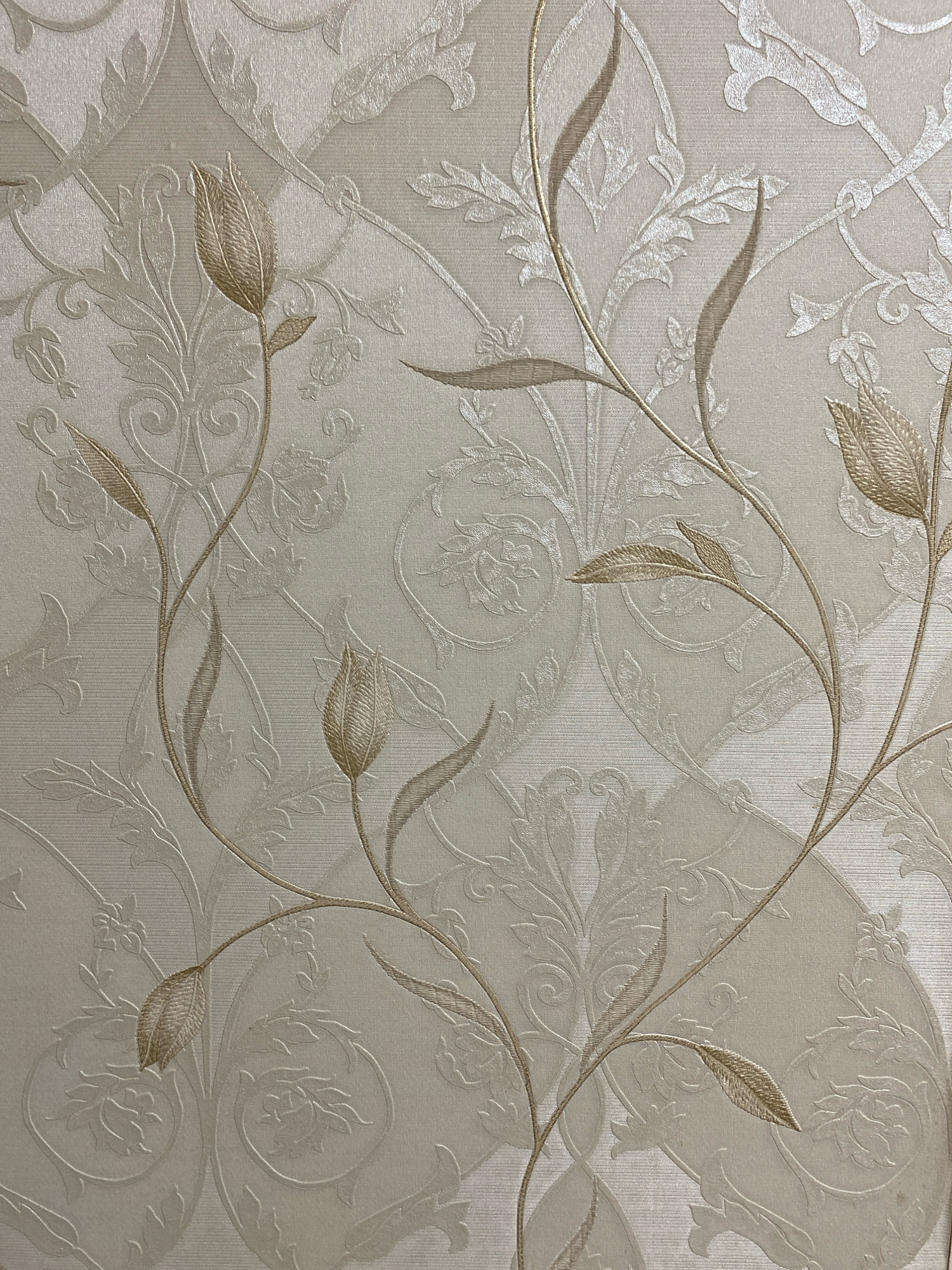 This is a close-up, top-down, eye-level, indoor, medium shot of a textured wallpaper. The wallpaper features a repeating damask pattern in light cream and beige tones. The raised pattern depicts stylized floral motifs and scrolling vines with leaves and unopened tulip-like buds. The overall impression is one of subtle elegance and vintage design. The lighting is even and diffused, highlighting the texture of the wallpaper. There are no people or external environmental elements visible in the image. The image is a still photograph and contains no information about the city of Ivanteyevka, Russia.