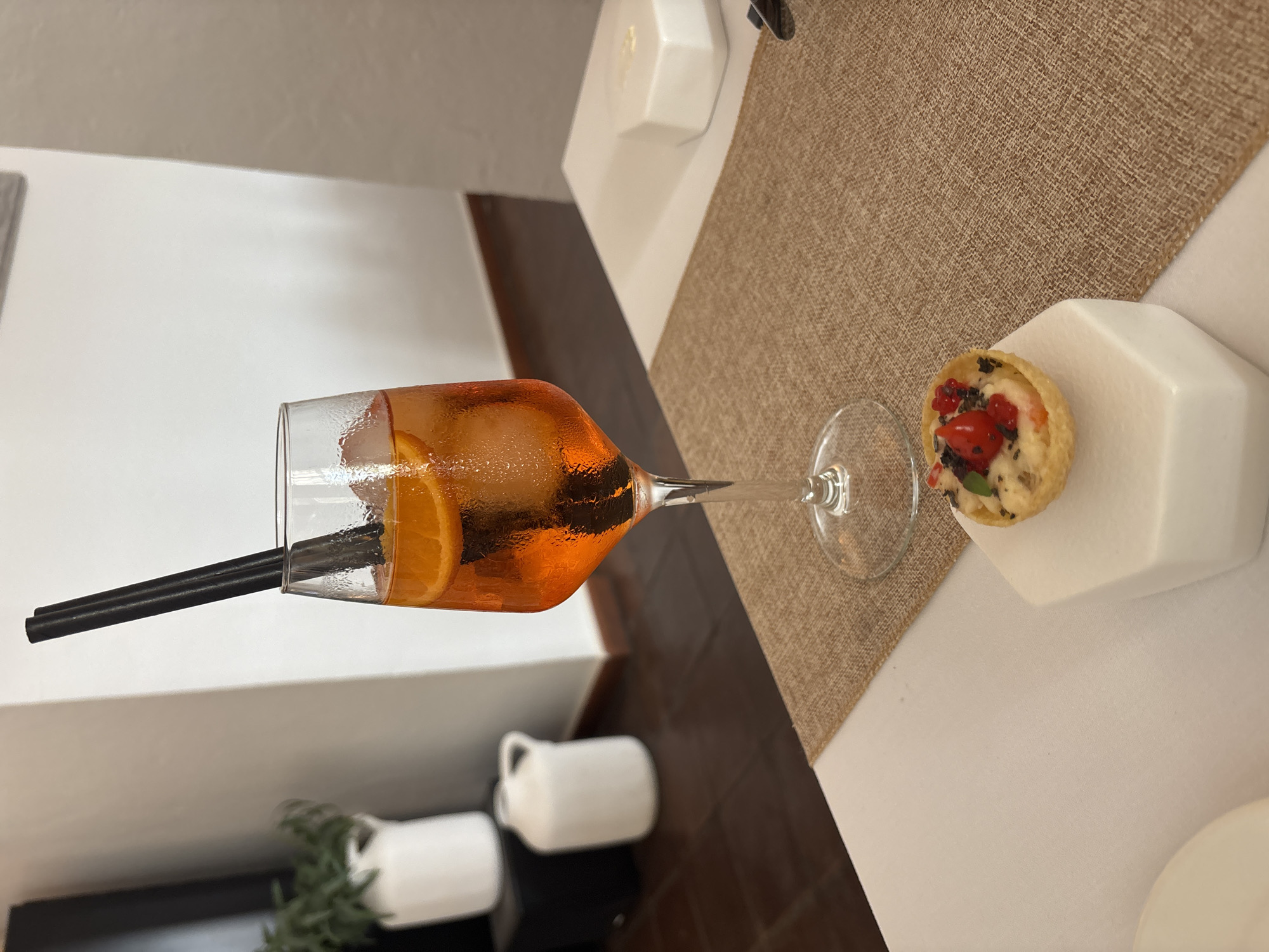 In Carboneras, Spain, on a table set with a white tablecloth and a brown burlap placemat, sits a tall, narrow glass filled with an orange aperitif and ice. Two black straws extend from the glass. In front of the glass, on a white geometric base, is a small tartlet topped with red berries, other small red and black garnishes, and a green leaf. In the background, a wooden floor is visible, along with white ceramic containers and a green plant in a white pot against a white wall. The scene appears to be indoors, likely a restaurant or bar. There are no people visible in the image.