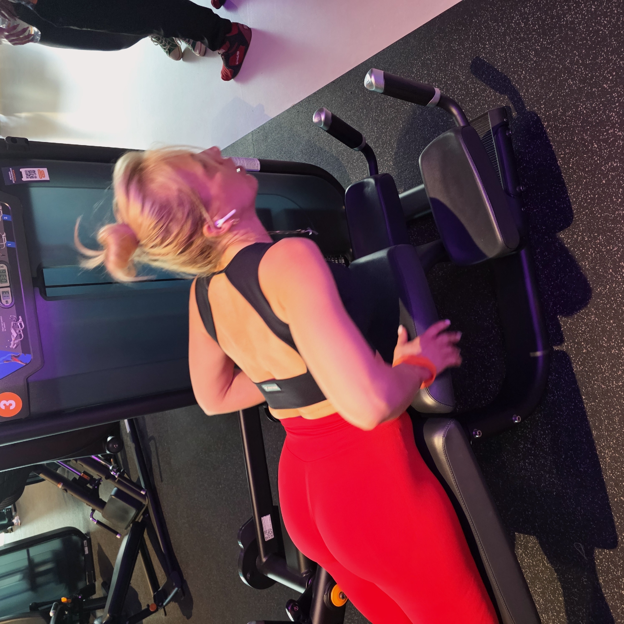 An angled, high-view shot captures a woman exercising in a gym in Saint Petersburg, Russia. The blonde woman, with her hair tied in a bun, is seen from a back-side perspective, focused on a piece of gym equipment. She wears a black sports bra with a cut-out design on the back, and vibrant red high-waisted leggings. White wireless earbuds are visible in her ears, and an orange wristband adorns her right wrist.
She is positioned on what appears to be a back extension machine or similar core exercise equipment, gripping handles on either side. The machine itself is dark-colored with padded support areas. In the background, another large piece of gym equipment features a screen displaying unreadable text and a clear orange circle with the number "3". A QR code is also visible on the side of this machine.
The gym setting features a dark, speckled rubber floor that meets a bright white wall. The scene is illuminated by strong artificial lighting, casting prominent shadows and creating a dynamic play of pink and purple hues across the subjects and equipment. Partially visible in the upper background are the legs and feet of another person, wearing dark pants and one red and black athletic shoe with unclear white text, and another shoe that is green and white. The overall atmosphere suggests an active, modern fitness environment.