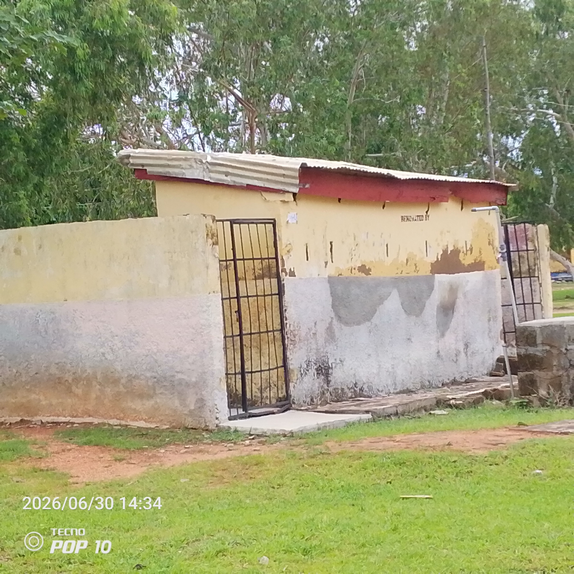 This image shows a building with a corrugated metal roof and peeling yellow paint. A black metal gate is visible on the left side of the building. The lower part of the building is rendered with a textured, off-white material. There are trees and foliage in the background, suggesting an outdoor setting. The ground in front of the building is covered with grass and some dirt. The overall scene appears to be somewhat run-down.
Visible text includes "REN OVATED BY" on the wall of the building and a date and time stamp "2026/06/30 14:34" along with "TECNO POP 10" at the bottom left corner. The time stamp indicates it was taken in the afternoon. The weather appears to be clear. The location context is Jalingo, Nigeria. There are no people visible in the image.