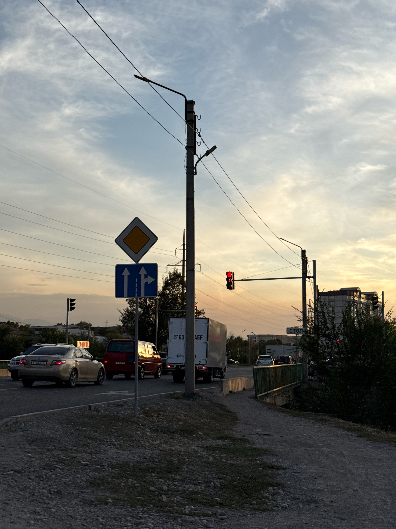 The image depicts a street intersection in Bishkek, Kyrgyzstan, during what appears to be the late afternoon or early evening, given the warm tones in the sky. A traffic light with a red signal is visible, indicating a stop. Road signs are present, including a diamond-shaped sign with a yellow center and a blue sign indicating lane directions. Several vehicles are on the road: a gold sedan, a red minivan, and a large white truck. Overhead power lines stretch across the frame, supported by utility poles. Buildings and trees form the background, with a bridge visible in the distance. The ground in the foreground is covered with gravel and some sparse vegetation. The overall impression is that of an urban street scene with active traffic flow.