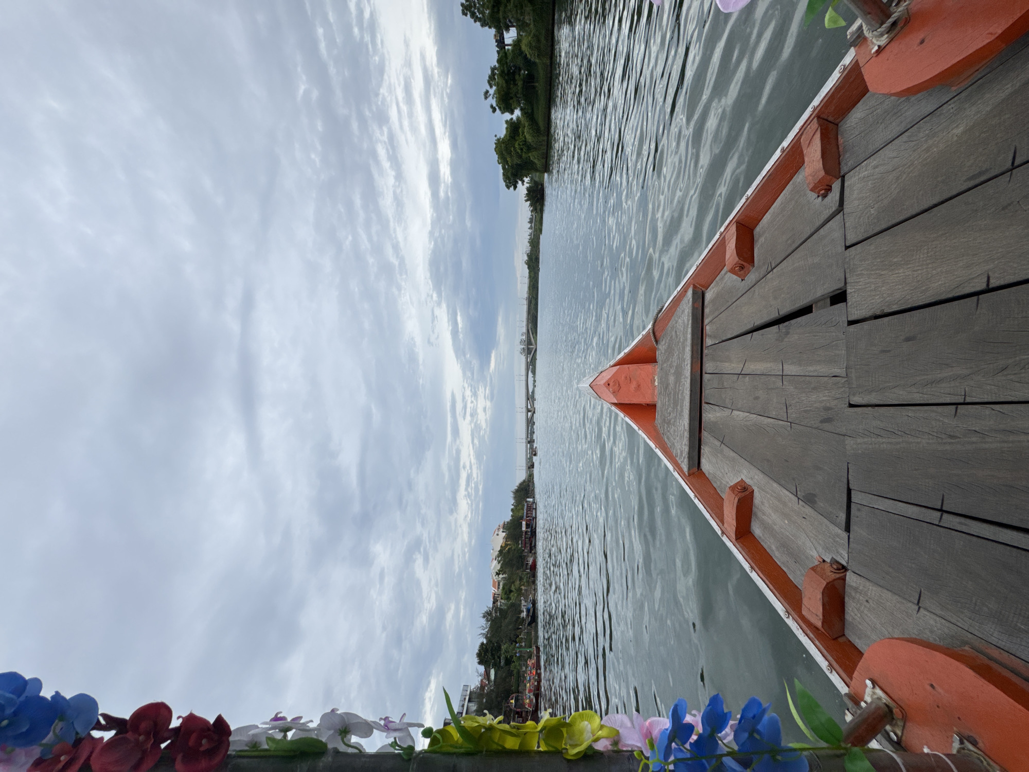 The image shows a view from the front of a boat traveling on a river, likely in the city of Đà Nẵng, Vietnam, given the presence of a bridge in the distance and lush vegetation along the riverbanks, typical of the region. The boat's wooden deck is visible in the foreground, with its orange-painted sides curving upwards. Artificial flowers in shades of blue, red, and white are attached to the left side of the boat, decorating the railing. The river's surface is rippled, reflecting the overcast sky. In the background, the river widens, with buildings and trees lining the shore. A bridge spans the river further in the distance, and the sky is filled with a layer of gray clouds. There are no people visible on the boat or in the immediate vicinity.