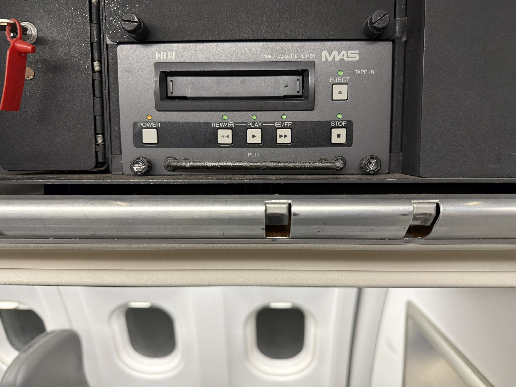 The image shows a Hi8 Video Cassette Player from MAS installed in what appears to be an airplane. The player has standard controls such as Power, Rewind/Play, Play/FF, Eject, and Stop. A handle labeled "PULL" is situated below the controls. The airplane interior, with its windows and seats, is visible in the background.